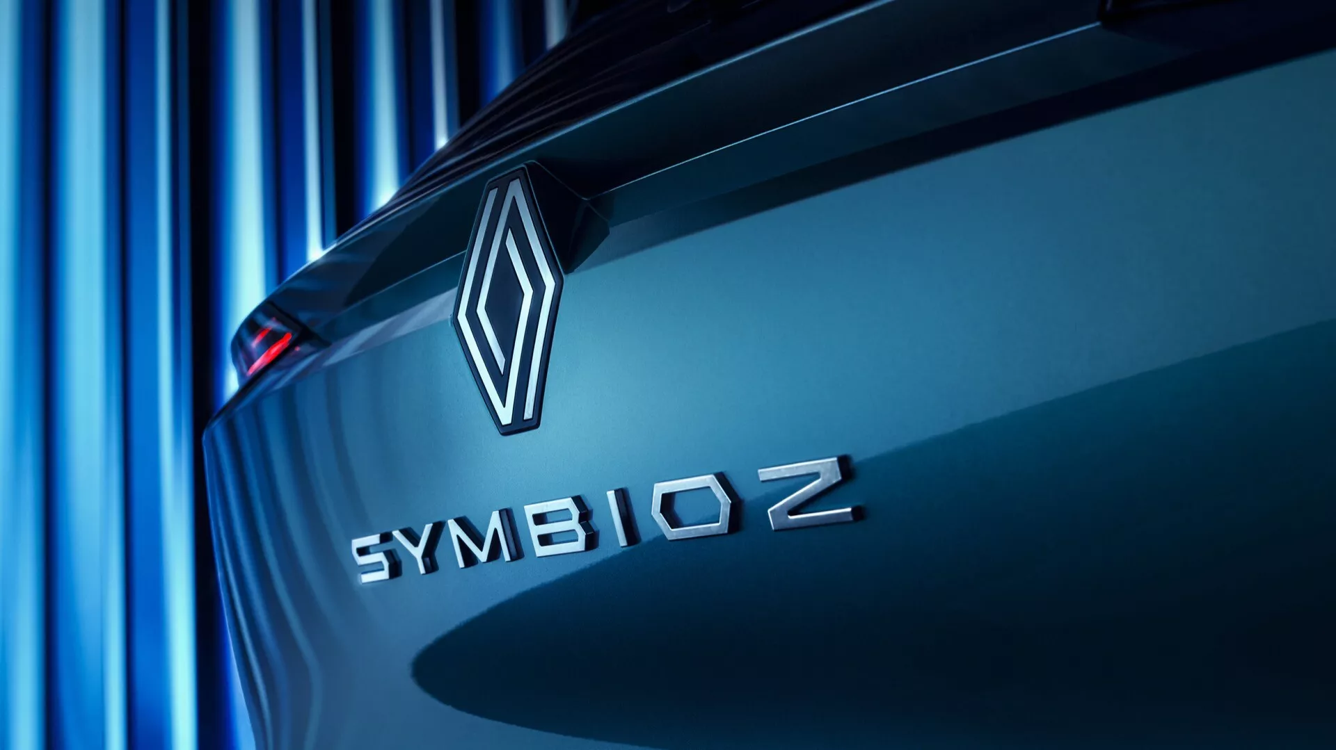 Närbild från ett vinklat perspektiv av bakre delen av Renault Symbioz 2024 (1. generationen), med fokus på varumärkesemblemet och modellnamnet. Diamantemblemet och 'SYMBIOZ' med versaler i sans-serif-typsnitt är tydligt synliga på bagageluckan, vilket indikerar en betoning på innovation och teknik. En mjuk blå ton kastar parallella linjer i bakgrunden och skapar en modern, elegant atmosfär. Bilen ser ut att vara stillastående, och vinkeln framhäver dess aerodynamiska linjer.