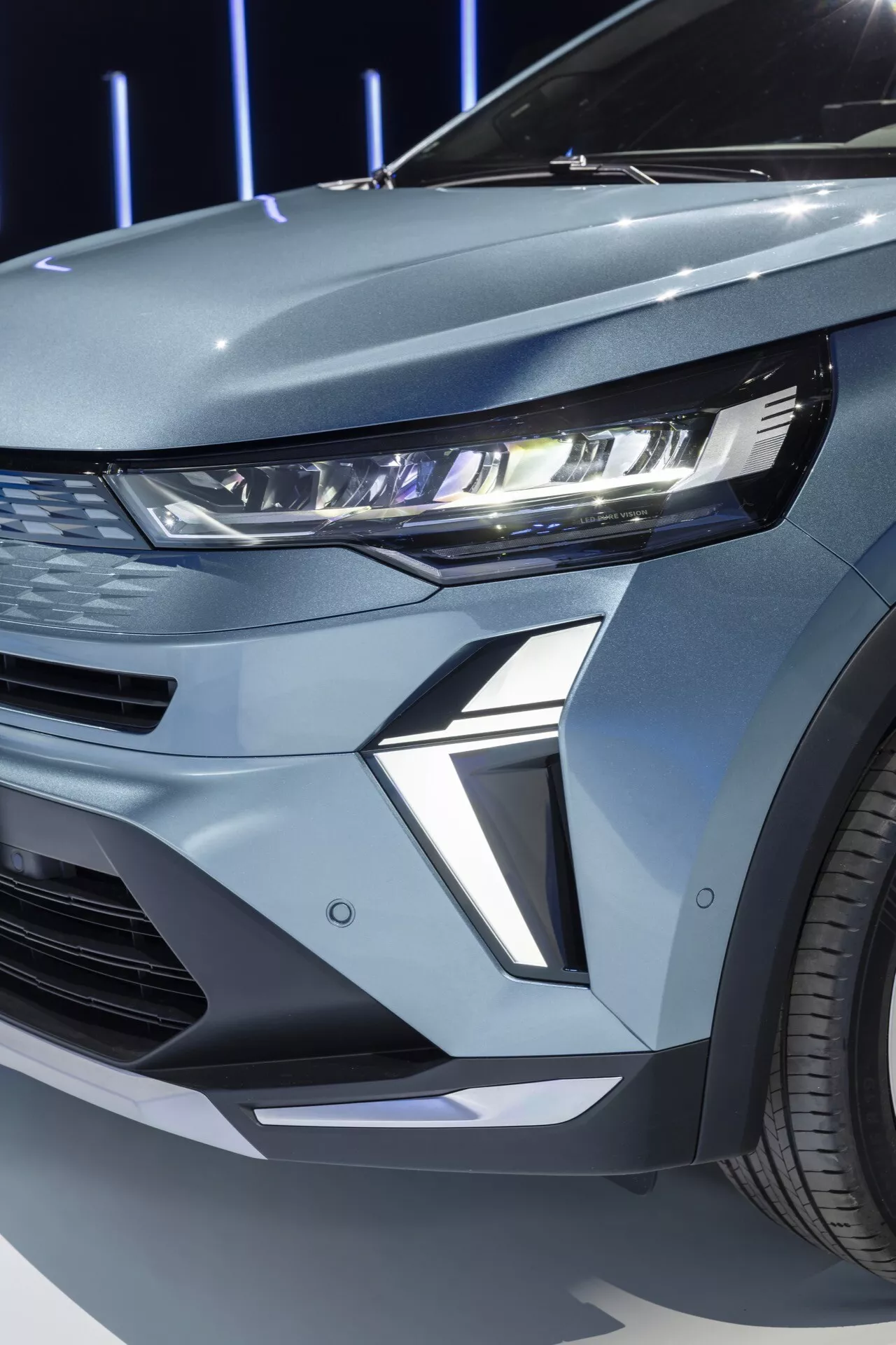 En närbild av den främre vänstra kvadranten av Renault Symbioz 2024 första generationen. Bilden visar fordonets moderna LED-strålkastare med en upplyst dagsljussignatur (DRL). Strålkastarhuset är smalt och sträcker sig in i skärmen, vilket exemplifierar samtida fordonsdesign. Den skulpterade sektionen under strålkastarna tillför en futuristisk och aggressiv look till bilen. Den synliga delen av den främre grillen antyder ett horisontellt intag eller en dekorativ panel som följer en sportig estetik. Bilens metalliska yta och den synliga flerspokesfälgen förstärker dess premiumkänsla.