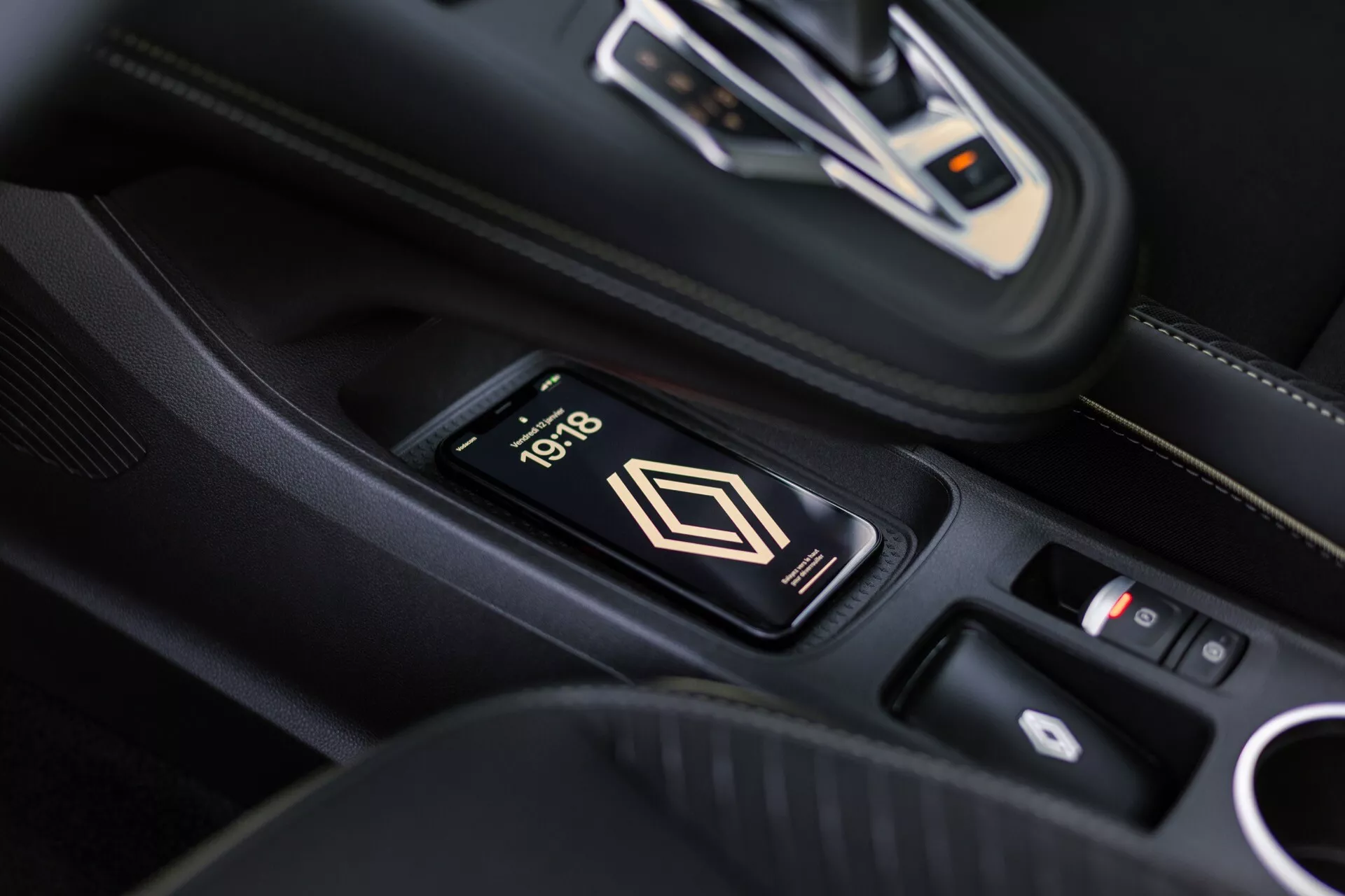 Interiörsbild av Renault Symbioz 2024s mittkonsol med en trådlös laddningsplatta för en smartphone som visar 10:18. Synliga är en elegant automatisk växelspak med metalliska accenter och en kontrollpanel i bakgrunden. Den lyxiga interiören har svart läder med vita sömmar.