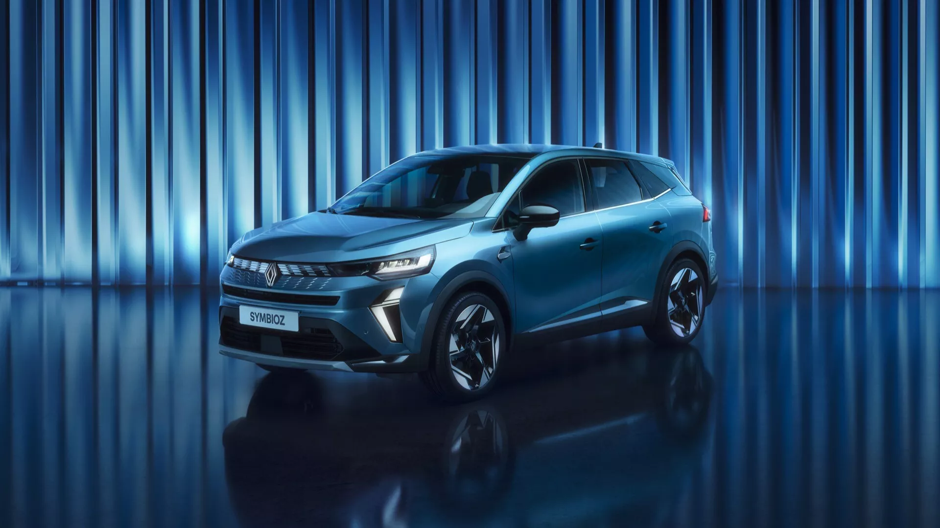 Bilden visar en Renault Symbioz 2024 (första generationen) SUV uppställd i en studio med en dynamisk blå bakgrund med vertikala ljusränder. En vinkel från låg till mitt visar bilens imponerande front och eleganta sidoprofil. Hela fordonet är i bildramen vilket betonar dess närvaro och moderna design. Synliga är skarpa, vinklade LED-strålkastare och en framträdande grill, tillsammans med en slät främre stötfångare som sannolikt rymmer säkerhetssensorer. Sidprofilen avslöjar aerodynamiska linjer, omlottiga dörrhandtag, strömlinjeformade sidobackspeglar och stora lättmetallhjul. Bilens metalliska finish och sportiga uppsyn är förstärkta av studiobelysningen och den reflekterande ytan under.