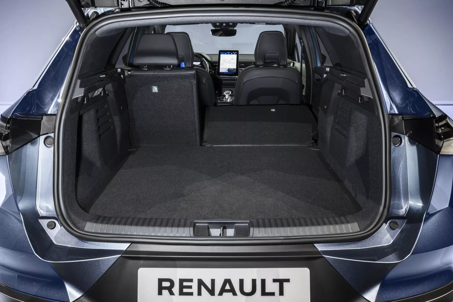 Vy in i lastutrymmet på en Renault Symbioz 2024 från en hög vinkel bakom den öppna bakluckan, som visar ett rent, oordnat bagageutrymme med upprätta säten, bakre stötfångare med Renault-märke, hållbar beklädnad, 12-voltsuttag på vänster sida och en krock på sidan fixa tillbehör på höger sida. Interiören och mittkonsolens skärm är synliga mot fronten, med jämn studioljus som framhäver funktionerna.