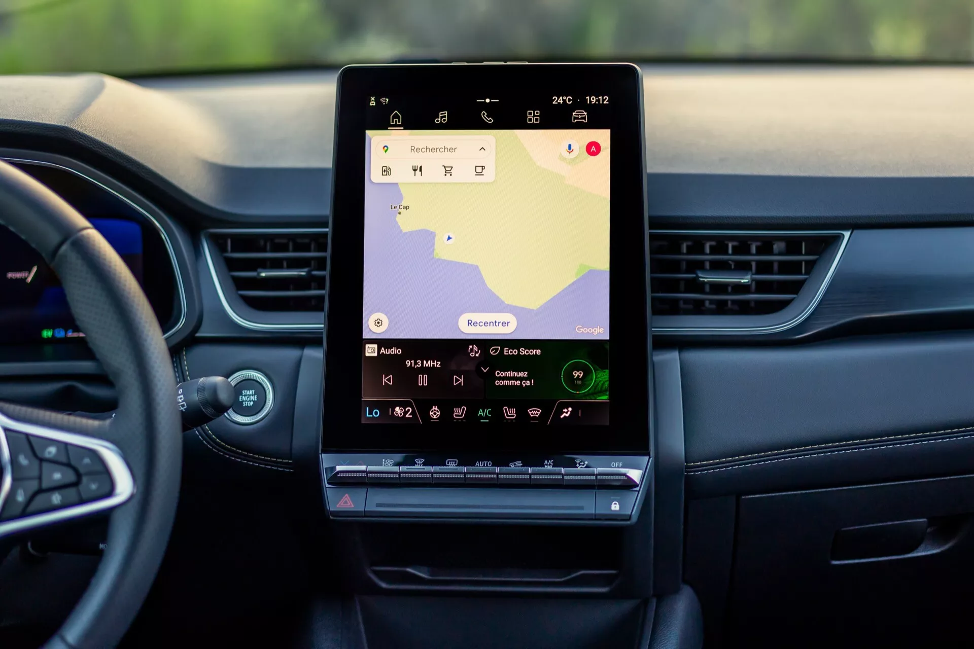 Interiörvy av Renault Symbioz 2024 (1:a generationen) från baksätets perspektiv, som visar en stor, vertikalt orienterad pekskärm på mittkonsolen med navigationskarta och multimediekontroller; modern instrumentbräda med horisontella linjer och luftventiler, delvis synligt ratt, som betonar avancerat infotainmentsystem och högteknologiska funktioner.