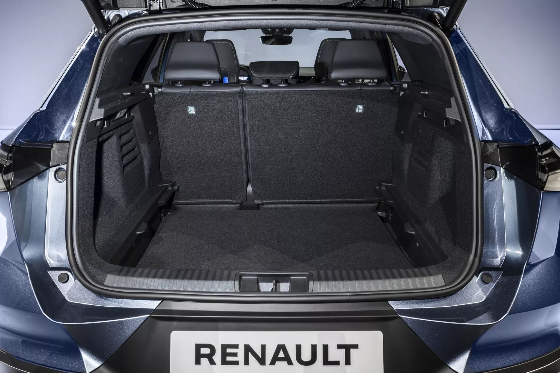 Interiörvy av Renault Symbioz 2024 lastutrymme från bakluckan, som visar ett symmetriskt bagageutrymme med svart matta, plastdetaljer och nedfällda baksäten för utökad förvaringskapacitet. Synlig märkning inkluderar 'RENAULT' under luckan.