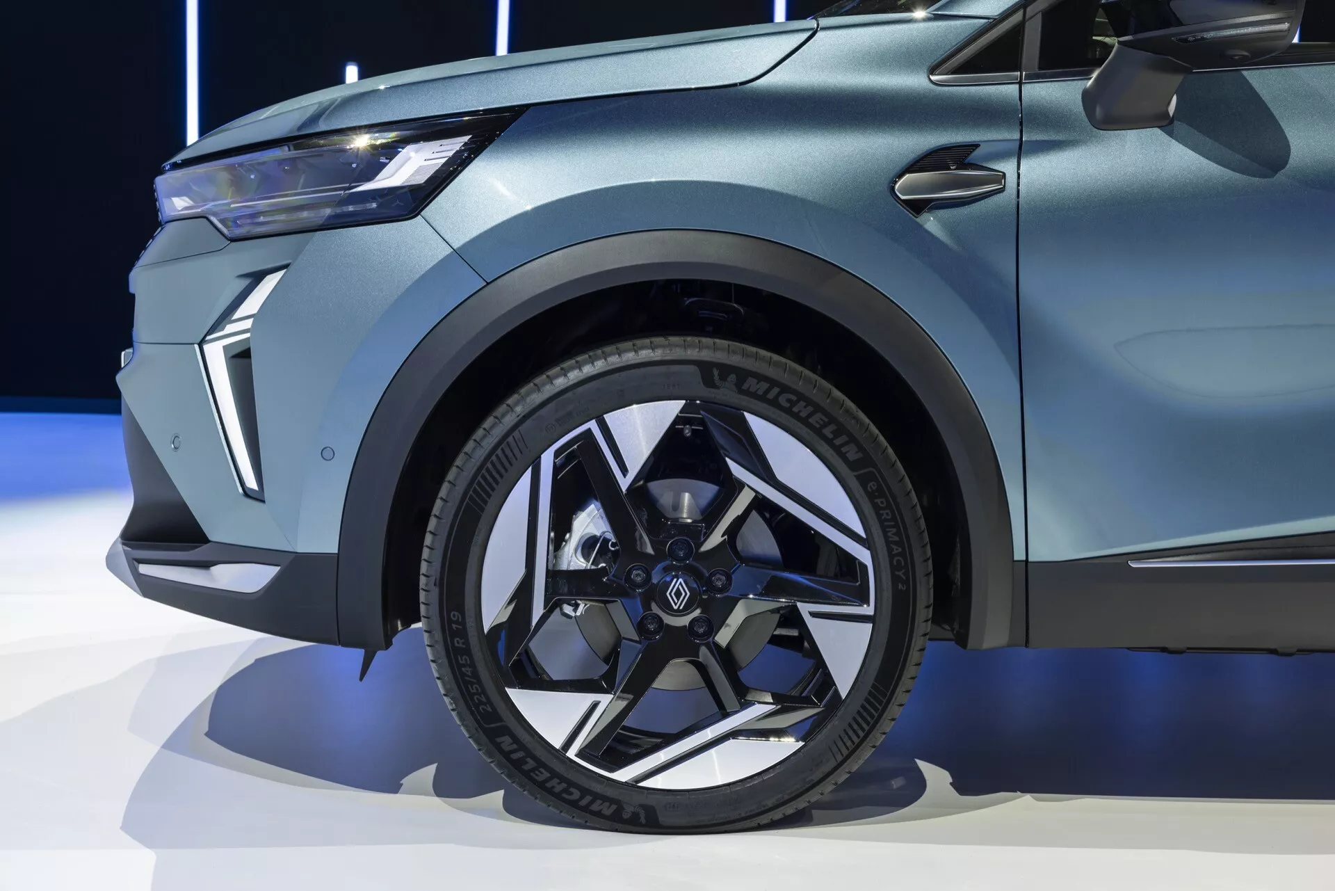 Närbild av den främre delen av en Renault Symbioz 2024 (1:a generationen), som framhäver det moderna svarta lättmetallhjulet med flerspakig design, lågprofildäcket med tydlig text på sidoväggen och de skarpa, muskulösa linjerna på skärmen. Bilden fångar också en liten del av dörren med en distinkt karosslinje samt den eleganta, vinklade ljusdesignen på den främre fasaden. Billackeringen verkar vara matt eller satin, vilket tyder på ett premium- eller specialutgåvemodell.