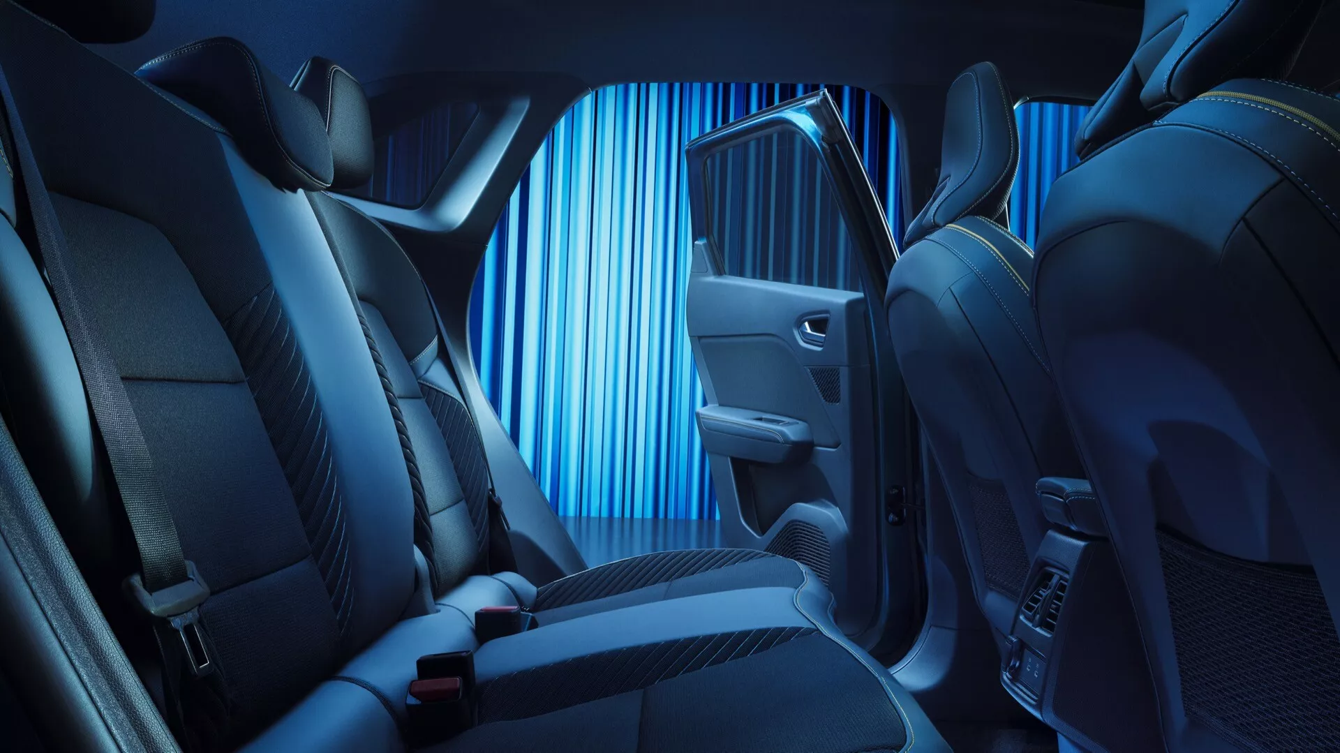 Bilden visar interiören av en Renault Symbioz 2024, med fokus på baksätesområdet från en vinkel inuti fordonets kupé, något mot passagerarsidan. Vyn är låg, nära sätesnivå, detaljerad med sätesmaterial, sömnader och design med blå ambient belysning som framhäver konturer. Synliga är baksätets layout med nackstöd och säkerhetsbälten, en del av framsätets passagerarsida, klädd med högkvalitativt tyg eller läder för lyx och komfort. Dörrpanelen är upplyst i blått och innehåller en kontrollknapp, möjligen för fönster eller andra avancerade funktioner, tillsammans med en högtalargaller och förvaringsfack. Den mörka mattan indikerar praktiskhet. Bilden antyder funktioner som avancerad ambient belysning, premium material, elektroniska kontroller och eventuellt ett premium ljudsystem, vilket återspeglar en fridfull och modern interiör fokuserad på passagerarkomfort.