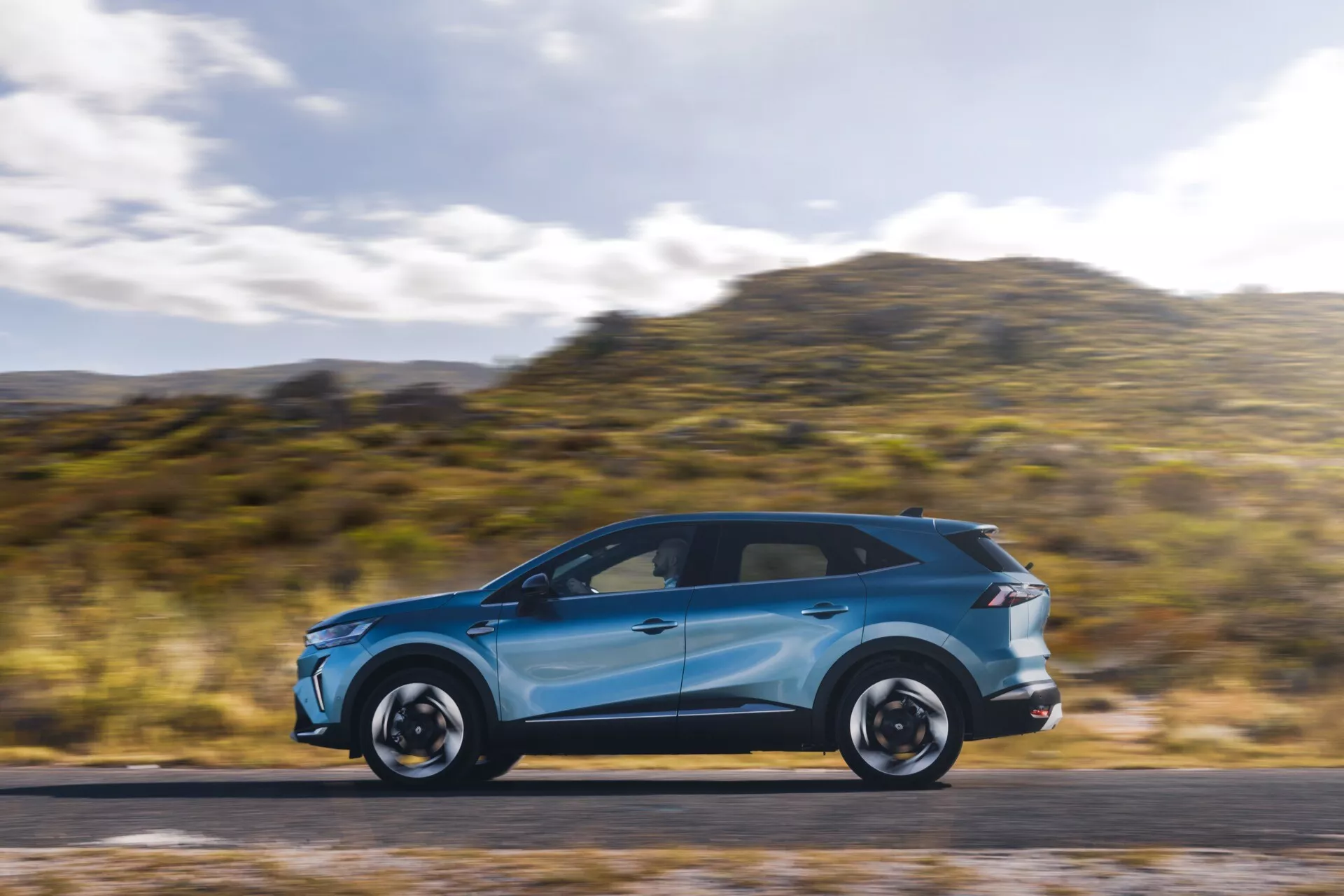 Ljusblå Renault Symbioz 2024 (1:a generationen) i rörelse, som visar sina eleganta aerodynamiska linjer och moderna design från en profilvy. Bilen, placerad i vänstra tredjedelen av bilden, har en suddig bakgrund och väg, vilket indikerar hastighet. Synliga funktioner inkluderar LED-dagsljus, integrerade blinkers i sidospeglarna, panoramasoltak, distinkta fälgar och lågprofildäck. Fotograferad med panoreringsteknik vilket framhäver bilens dynamiska rörelse.