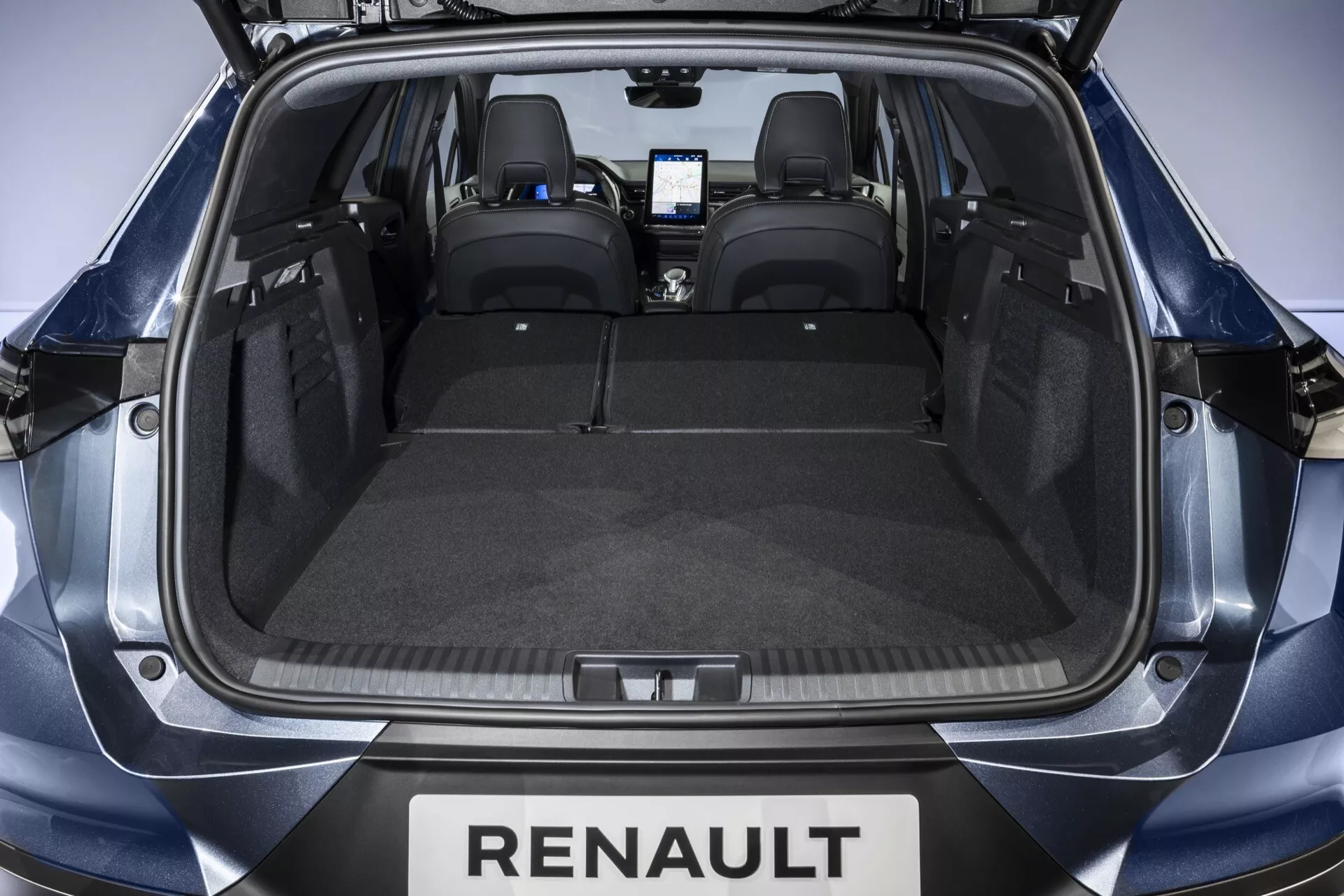 Vy från baksidan med öppen baklucka in i lastutrymmet på Renault Symbioz 2024. Det rymliga bagageutrymmet har ett platt golv täckt med grå matta, designat utan hinder för att maximera kapaciteten. Två spärrar på varje sida antyder att baksätena kan fällas ner för mer utrymme. De svarta läderbaksätena med nackstöd och det svagt synliga toppmoderna infotainmentsystemet i frontkabinen antyder en teknikfylld interiör.