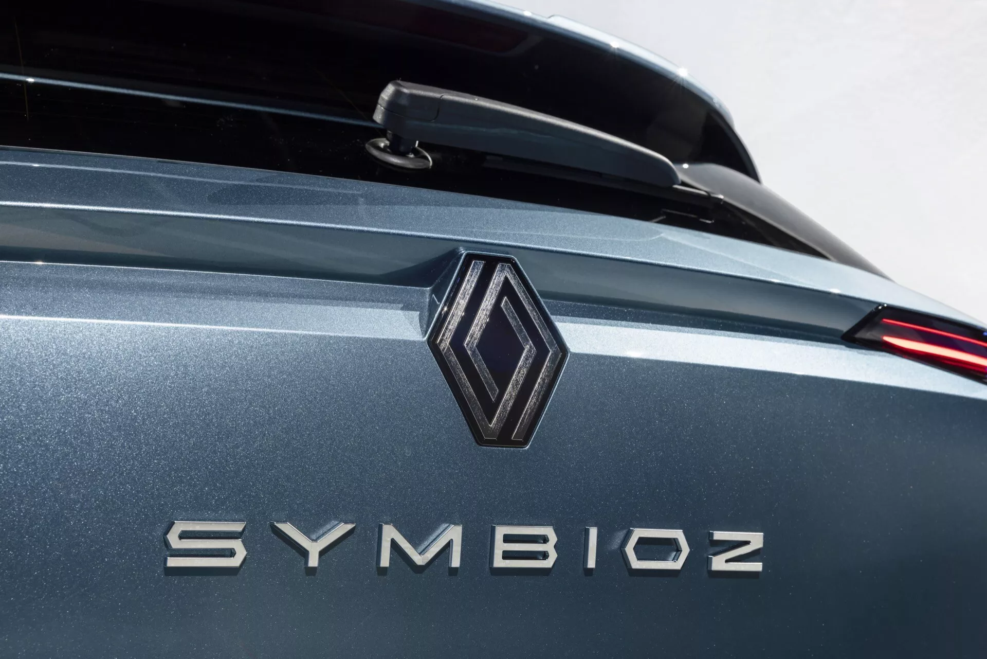 Professionell närbild av bakre delen av en bil med fokus på märkesdetaljer med en diamantformad emblem och modellnamnet 'SYMBIOZ' i metalliska bokstäver. Bilden använder ett litet skärpedjup som framhäver emblemet och bokstäverna medan bakgrunden mjukt suddas ut. En väl upplyst miljö antyder en inomhusutställning eller studio. Fordonets färg är en metallisk grå eller blå nyans med reflexiva egenskaper, och har eventuellt en bakre spoiler och modern design med LED-bakljus.