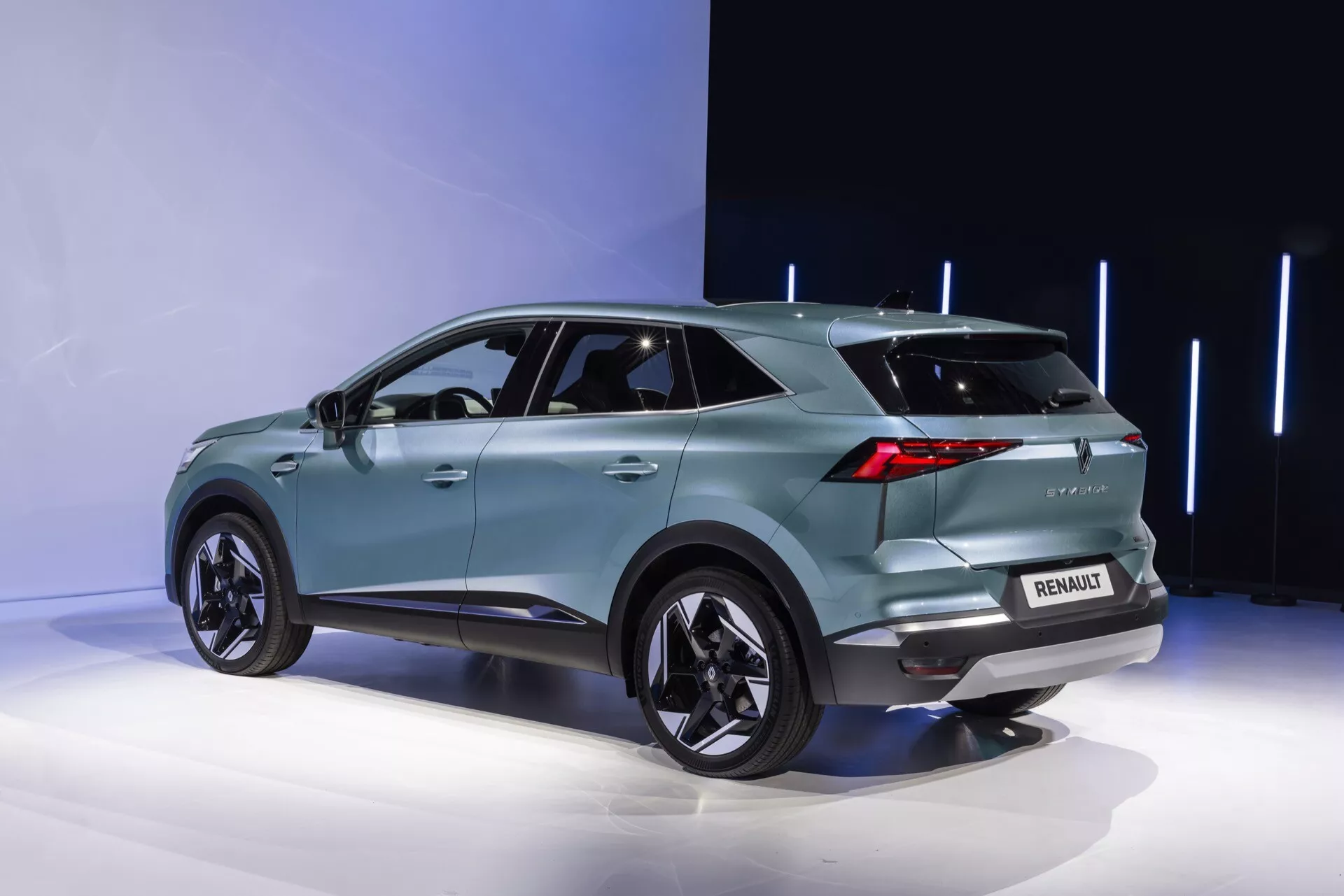 Trekvart bakifrån av Renault Symbioz 2024. Den turkosa SUV:en, placerad på en visningsplattform, visar upp en modern aerodynamisk design med sluttande taklinje, uttalade hjulhus med stora fälgar och tonade bakrutor. Distinkt LED-bakljus, en bakre spoiler, synligt varumärke med logotyp och modellbeteckning, parkeringssensorer och eventuellt en backkamera är synliga. Den eleganta bakre stötfångaren har också ljuselement, medan en hajfensantenn antyder avancerad teknik. Integrerade blinkers i ytterbackspeglarna och infällda dörrhandtag tyder på nyckelfri åtkomst. Den minimalistiska bakgrunden framhäver fordonets detaljer.
