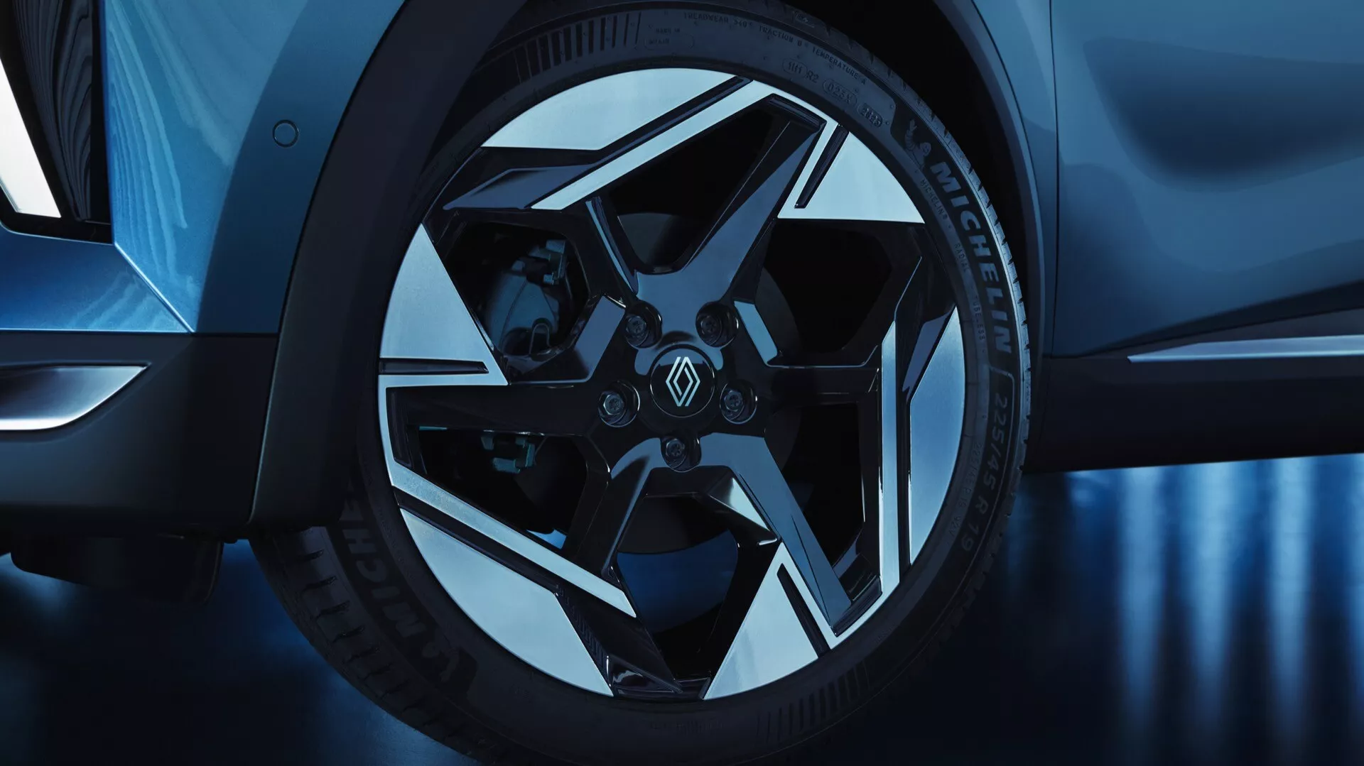 Närbild av framhjulet och delvis sidoprofil av Renault Symbioz 2024 (1:a generationen) tagen från en låg vinkel. Aluminiumhjulet har en komplex, geometrisk design med flera ekrar och växelvis glansiga och matta ytor, i kombination med lågprofilerade prestandadäck och märkesbromsok, vilket antyder sportighet och lyx. Den kalla blå tonen förstärker estetiken och antyder studiobelysning eller efterbearbetning.
