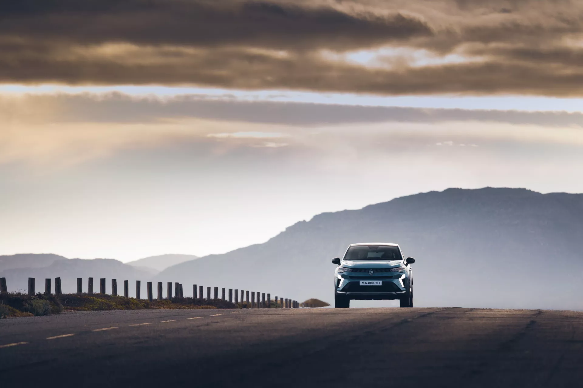 Frontal vy av Renault Symbioz 2024 (1:a generationen), fångad under tidig morgon eller sen eftermiddag med varmt ljus och långa skuggor. Fordonet, med eleganta designlinjer och framträdande strålkastare, rör sig mot kameran på en hög höjdfartväg, med en lätt oskarp bakgrund av berg. Ett upplyst molnigt himmel tillför en atmosfärisk kontext; den låga perspektiven betonar frontfunktioner som grillen, huven och stötfångaren. Rörelseoskärpa vid kanterna illustrerar bilen i rörelse samtidigt som den behåller skarp fokus på modern teknik och design.