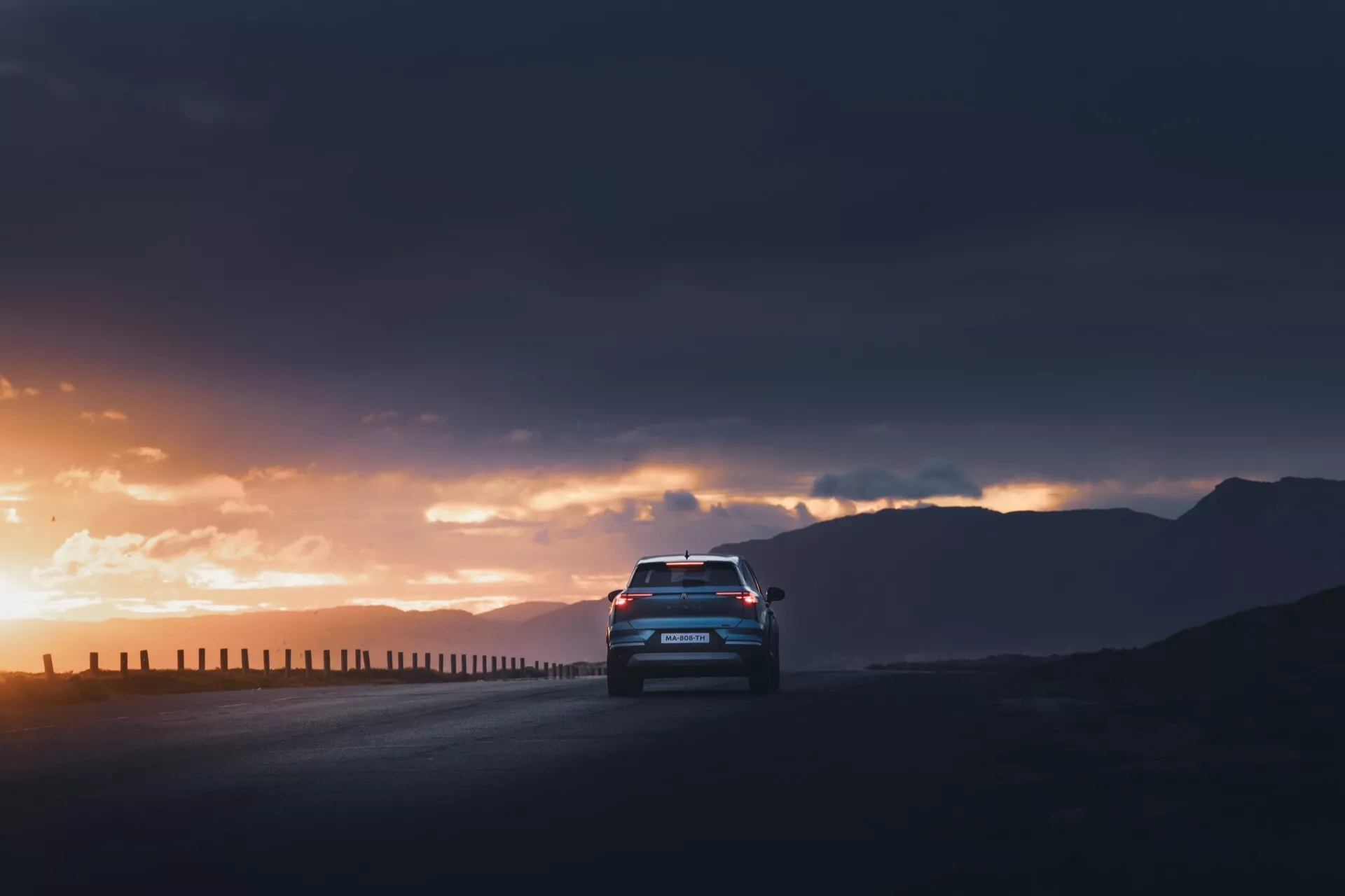 Låg vinkel bakifrån av en Renault Symbioz 2024, 1:a generationen, som kör på en väg mot en dramatisk soluppgång eller solnedgång. Bilens silhuett framhävs av upplysta LED-bakljus och bakre designdetaljer inklusive en spoiler, dubbla avgasrör och en bakre diffusor. Den mörka, molniga himlen med solstrålar som bryter igenom molnen förstärker bilens närvaro, medan reflexerna på bakrutan antyder en inre belysning eller ambient belysning.