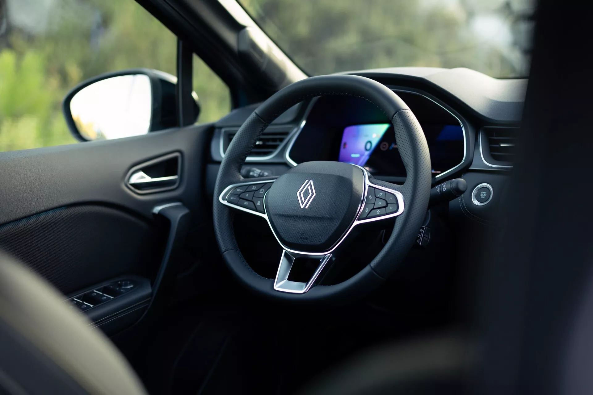 Interiörvy av Renault Symbioz 2024 från baksätet, med fokus på den moderna treekrade ratten och den digitala instrumentpanelen. Ratten har en platt botten, metallisk accent och integrerade kontrollknappar, omgiven av ett mörkt färgschema. Den grunda skärpedjupet framhäver dessa element, med oskärpa i omgivningen inklusive ett nackstöd och utomhusscen genom fönstret, vilket antyder att bilen står stilla.