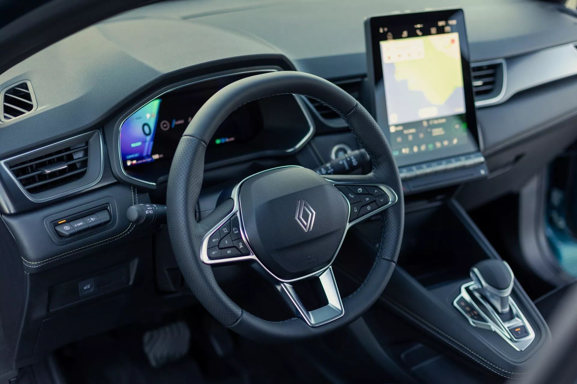 Interiörbild av Renault Symbioz 2024 (1:a generationen) från förarens perspektiv, fokuserad på ratt och instrumentbräda. Bilden ger en fågelperspektiv, 30-graders nedåtvinkel som visar en treekrad ratt med multifunktionsknappar och ett generiskt emblem, en helt digital instrumentpanel och ett stort pekskärmsinfotainmentsystem som visar en karta. Mittkonsolen har en modern växelväljare med minimalistisk design. Interiören visar en högteknologisk design av elbil med ambientbelysning och modern estetik.