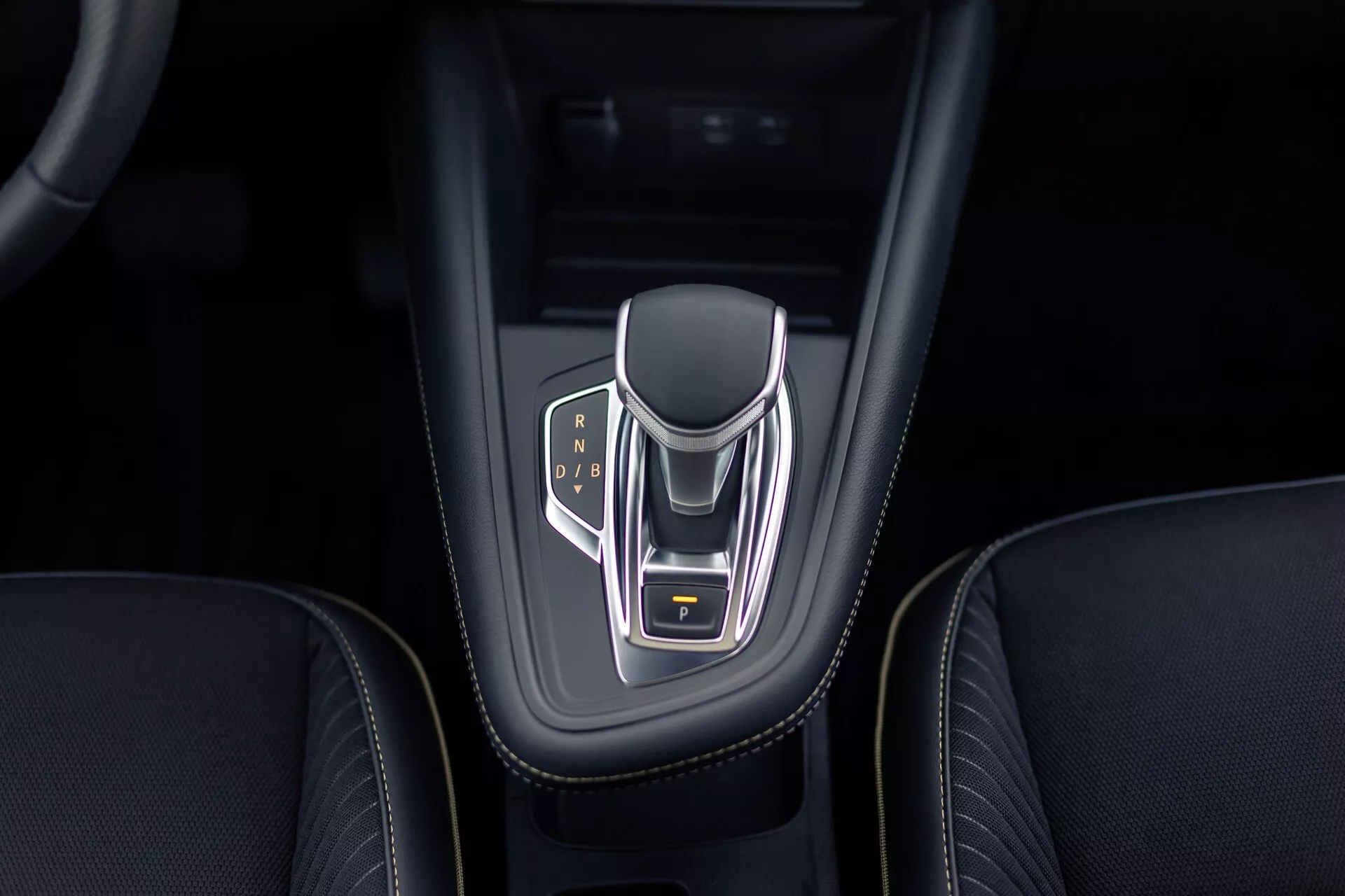 Vy från ovan på Renault Symbioz 2024 mittkonsol med fokus på den elektroniska växelväljaren med kromfinish och belysta växelindikatorlampor för Park, Reverse, Neutral och Drive. Växelväljaren har sidor belagda i läder, en gulmarkerad parkeringsknapp och är placerad i ett glansigt svart hölje med intrikata sömmar på den omgivande läderinteriören.