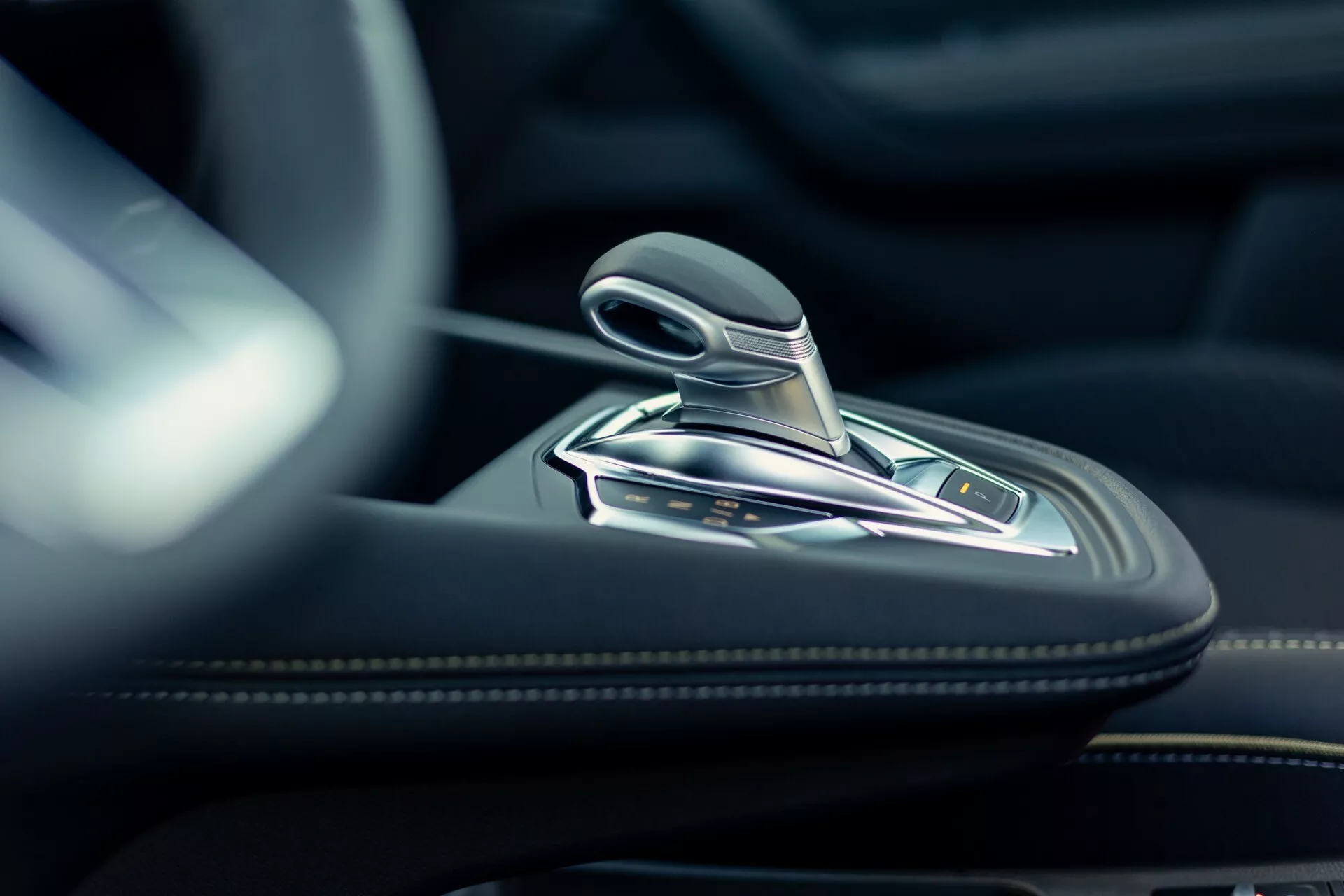 Närbild av interiören i Renault Symbioz 2024 som visar en modern växelspak med metalliska detaljer och troligtvis en läderfinish. Högupplöst bild tagen från låg vinkel som fokuserar på den automatiska växelväljaren och den omgivande mittkonsolen, tillverkad av högkvalitativa material som läder, borstad metall eller krom. Flera knappar som troligen styr körlägen, parkeringsbroms, auto-hold och traction control är synliga på konsolen. Mjukt, naturligt ljus framhäver bilens lyxiga och precisa interiördesign.