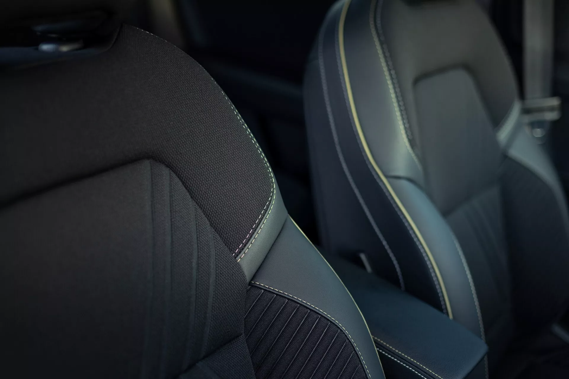 Närbild av framsätet på passagerarsidan i Renault Symbioz 2024 (1:a generationen). Sätet har en mittpanel av texturerat tyg eller syntetisk väv för bättre andningsförmåga, omgiven av slät, eventuellt syntetiskt läderliknande material med kontrasterande dekorativa sömmar, vilket framhäver dess premium- eller sportiga design. Det grunda skärpedjupet fokuserar på sätet och suddar ut bakgrunden för att betona materialkvaliteten och hantverket.