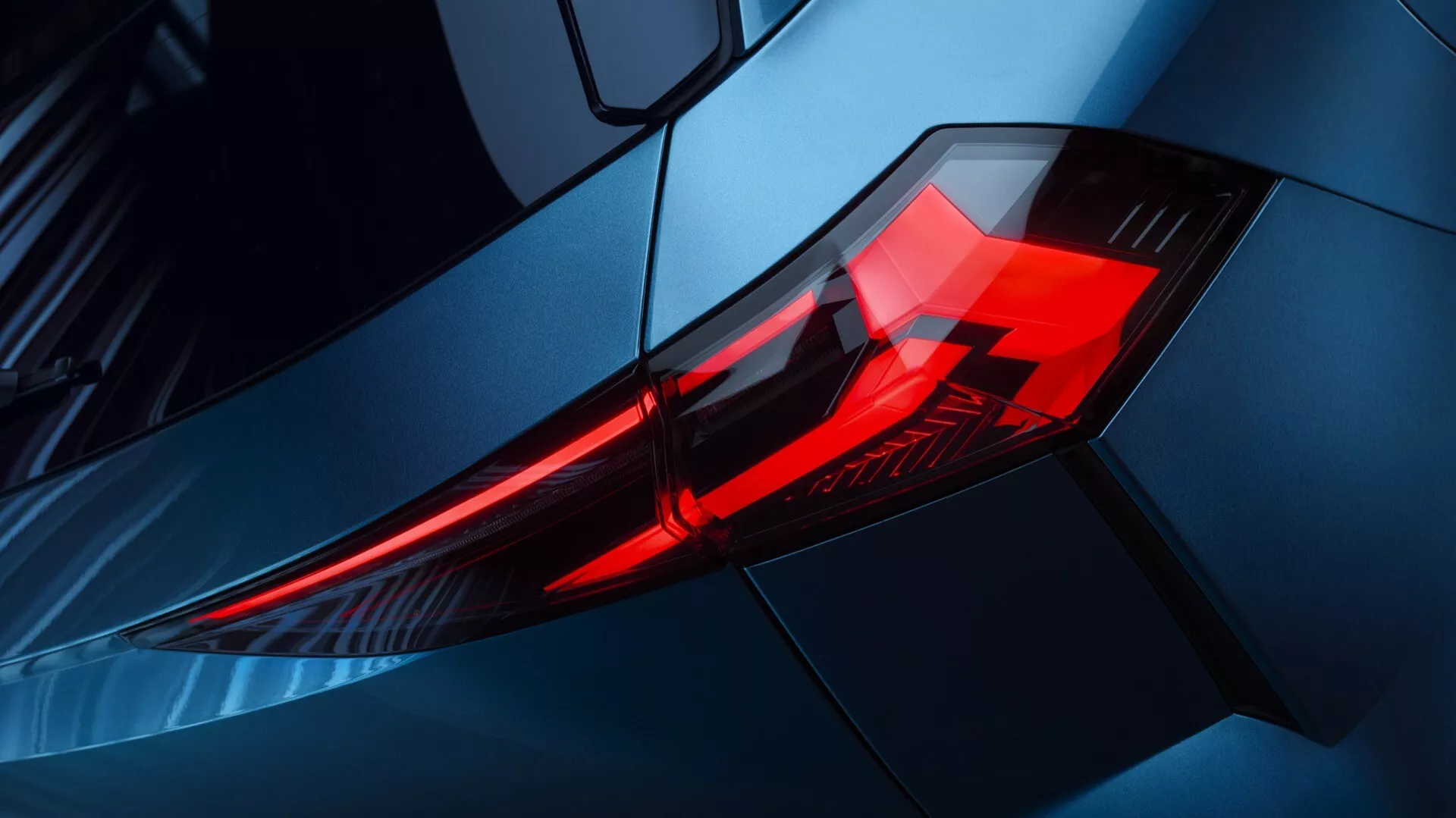 Närbild av baklyktan på Renault Symbioz 2024, som visar LED-belysningselement i en vinklad, lagerdesignad hölje. Fotot är taget från en sned vinkel, fokuserat på det bakre vänstra hörnet med en upphöjd synvinkel, som framhäver bilens metallicblåa kaross och aerodynamiska linjer. Bilden betonar fordonets moderna, sportiga design och premiumestetik, lämplig för diskussioner om fordons- och designtrender.
