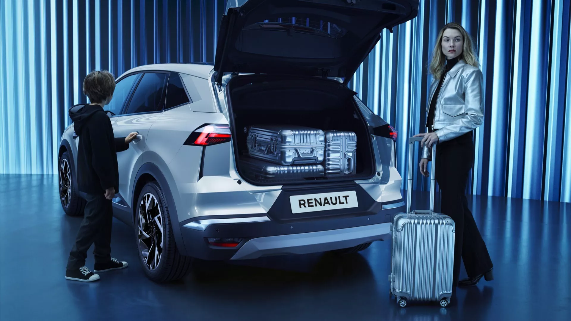 Trebaks vy av en vit 2024 Renault Symbioz SUV i en showroommiljö, med öppen baklucka som visar det rymliga bagageutrymmet, moderna LED-bakljus, Renault-logotypen under bakrutan, parkeringssensorn på stötfångaren, flerspårslegerade hjul, tryckknappsautomatisk öppningssystem, och två personer: en kvinna med resväska som antyder resa och ett barn som interagerar med bagageutrymmet och framhäver fordonets familjevänliga egenskaper.