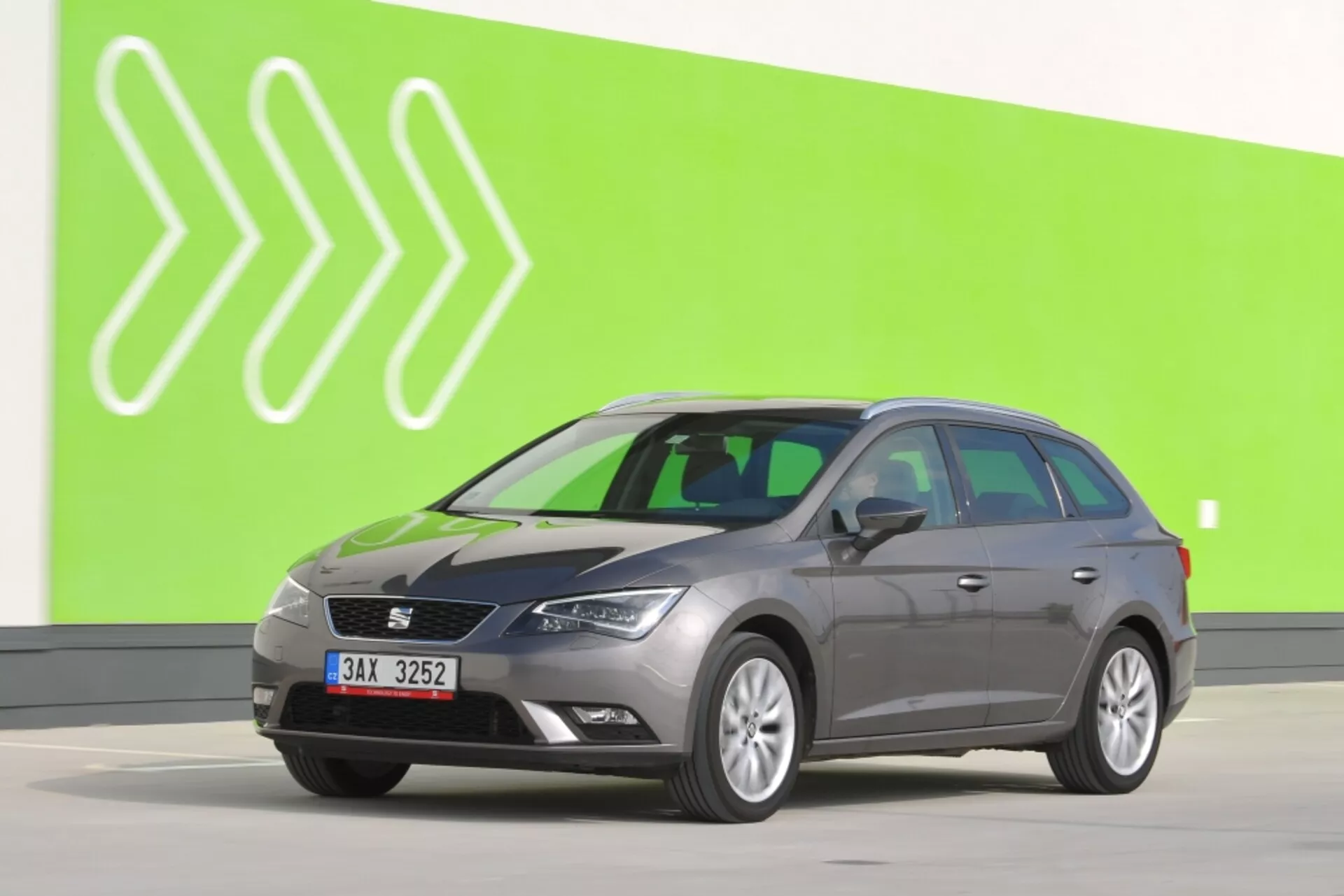 Grå Seat Leon SC 2012 station vagn i full sidovy mot en grön bakgrund med stiliserade vita pilspetsar, visar sidospeglar med svängsignaler, karakteristiska dörrkonturer och standardhjul på en ljusgrå studiovåning