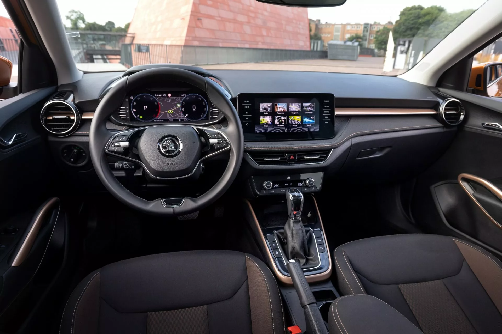 Interiör av Skoda Fabia 2021 från baksätet, visar en treekrad ratt med kontroller, digital instrumentpanel som visar hastighetsmätare och varvräknare, och central pekskärmsinfotainmentsystem med ikoner för navigation och musik. Runda och horisontella luftventiler med trä- eller metallimiterande finish, automatväxel med elektronisk parkeringsbroms på mittkonsolen, sportiga framsäten i läder med kontrasterande sömmar. Modern atmosfär och rymligt inre som indikerar lyx och sportighet.