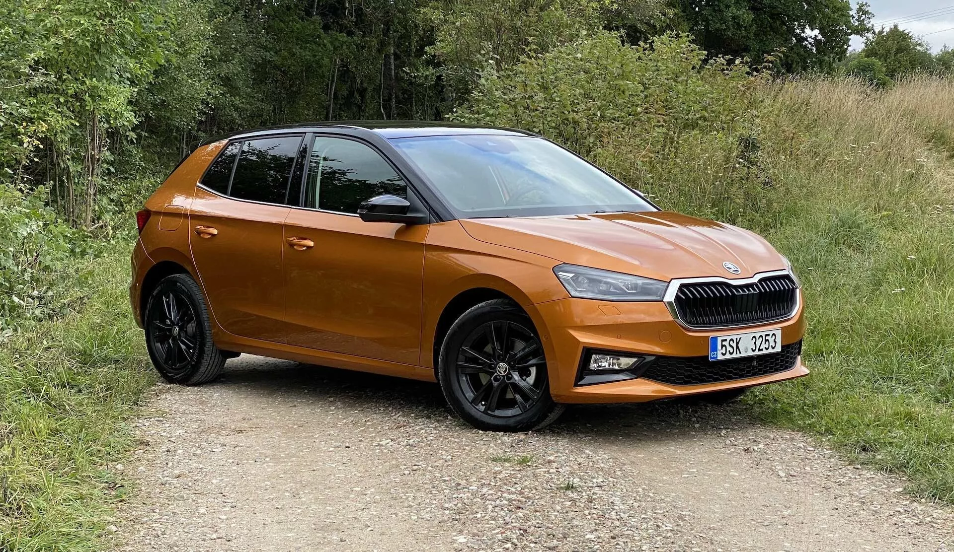 Bild av en modern Škoda Fabia 2021 hatchback, tagen från en trekvartsvy framifrån, som visar vänstra framsidan, förarsidans dörr, vänstra sidan av motorhuven och en del av taket. Bilen är placerad på en grusväg omgiven av grönska, vilket indikerar en lantlig miljö. Synliga funktioner inkluderar en horisontellt slätad grill med centralt emblem, skarpa LED-strålkastare, metallisk orange lack, sidospeglar i samma färg som karossen, svarta flerspoked fälgar med mönstrade däck och konturerade dörrar med aerodynamiska linjer. Bilen verkar vara ny, kompakt och utan modifieringar.