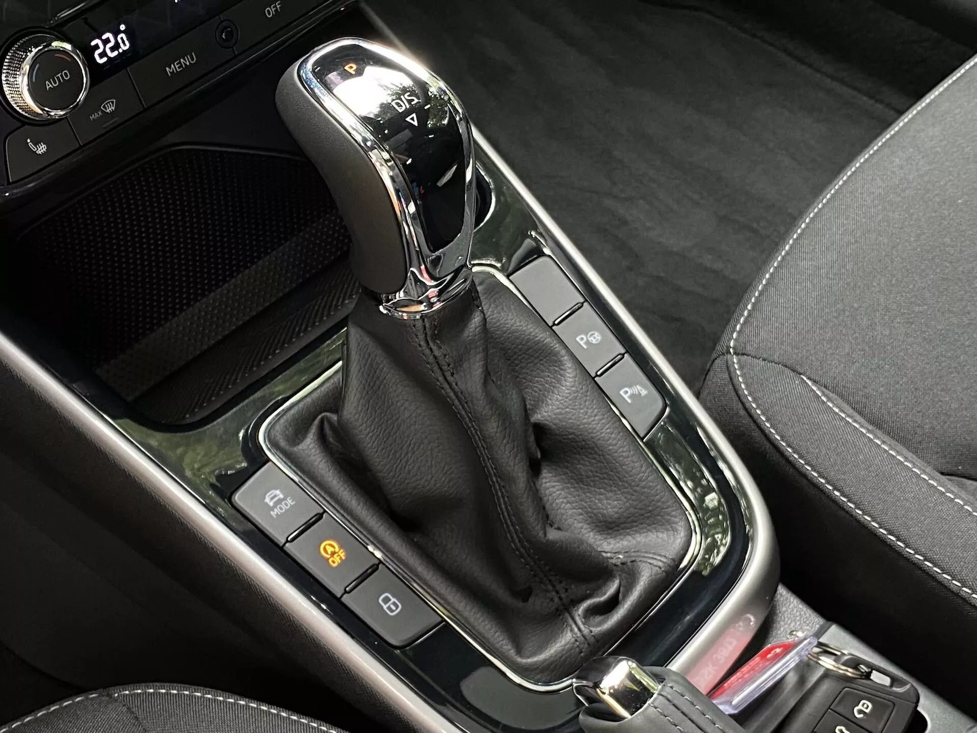 Närbild av den automatiska växelspaken i mittkonsolen på en Skoda Fabia 2021. Den moderna växelspaken, placerad vertikalt i 'P'-läge (Parkering), har en krom- eller metallfinish på ovansidan och är innesluten i en blank svart panel. Under växelspaken finns en svart läderstövel som döljer mekanismen. Runt växelspaken finns en mängd knappar och indikatorer, inklusive två till vänster som troligen är relaterade till bilens säkerhetsfunktioner, och en del av en röd knapp till höger. Området kombinerar olika svarta material, inklusive blanka ytor och mönster som imiterar kolfiber, vilket antyder en modern högteknologisk design.