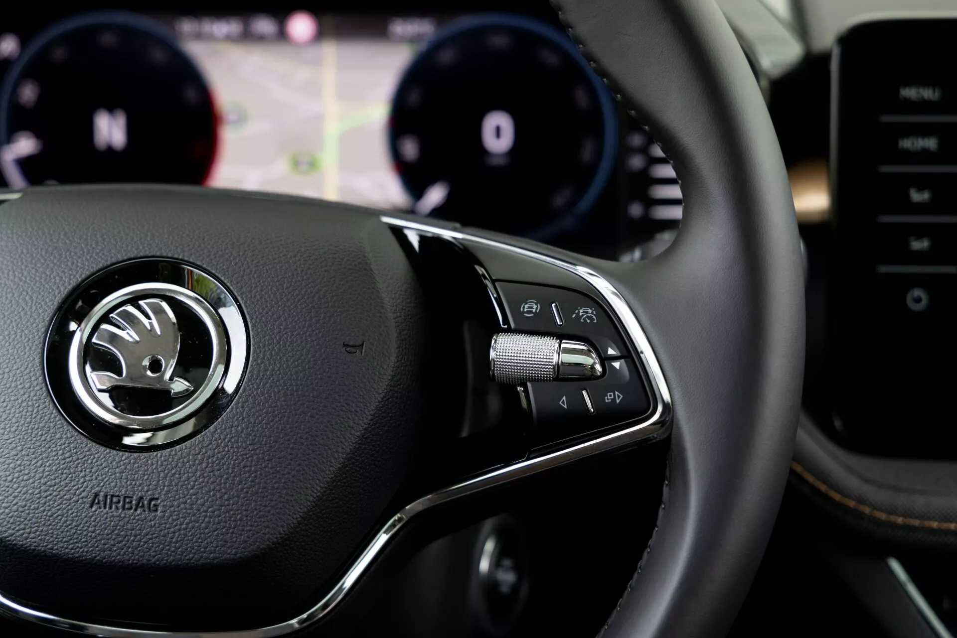 Närbild av interiören i 2021 års Skoda Fabia från förarens perspektiv, med fokus på de högersidiga kontrollerna på ratten. Den läderklädda ratten har ett centralt silveremblem med Skodas logotyp och texten 'AIRBAG' präglad under. Högra kontrollpanelen inkluderar knappar för infotainmentsystemet, handsfree-samtal eller röstkommando, med paddlar synliga bakom ratten. Den suddiga bakgrunden visar instrumentklustret med varvräknare och hastighetsmätare samt en digital display för förarassistans. Central konsol eller multimedia system delvis synlig på höger sida.