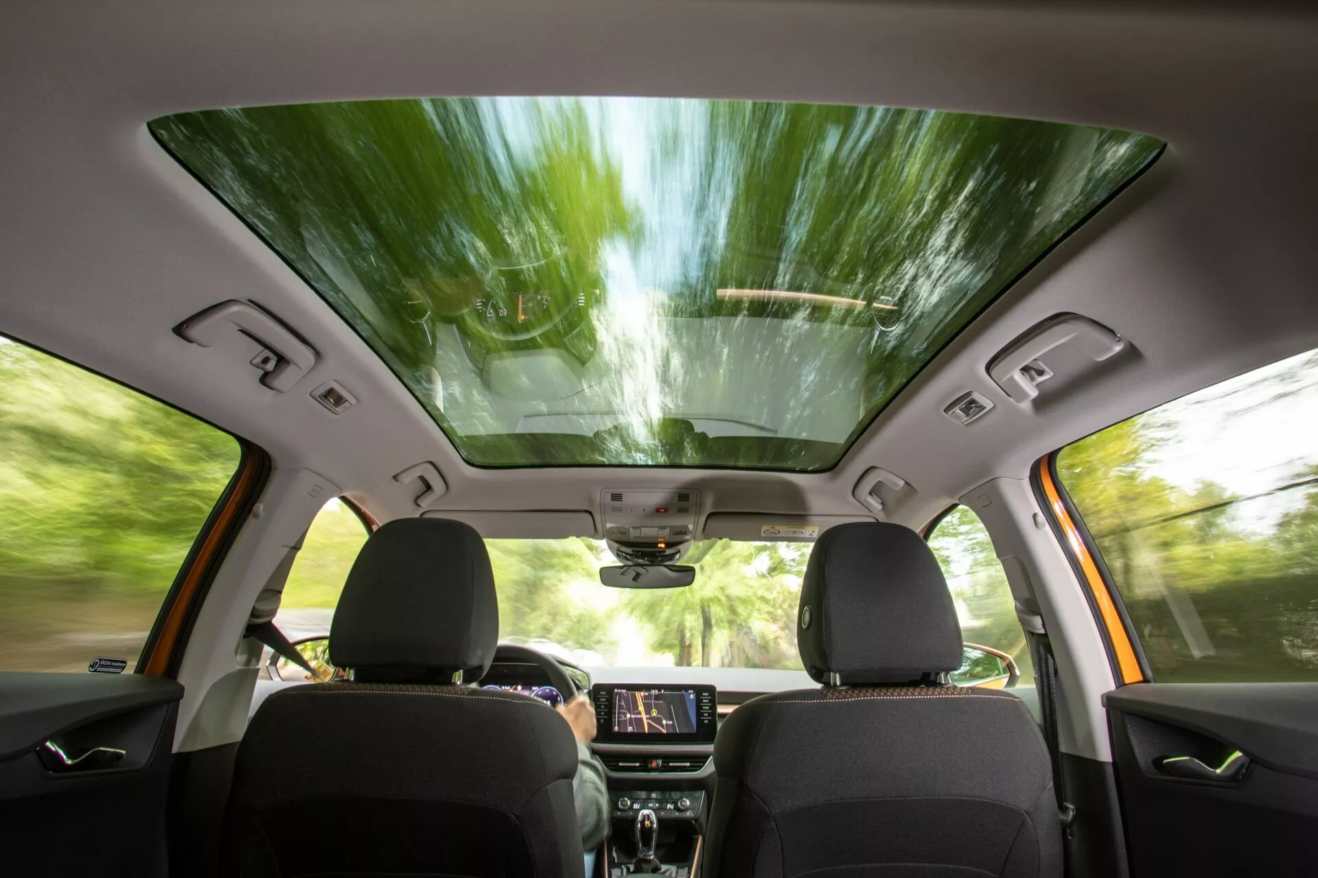 Interiörvy av 2021 års Skoda Fabia från baksätets perspektiv, som visar framsäten med mörk klädsel, panoramatak, upplyst infotainment-pekskärm på instrumentpanelen, kontrollpaneler, dörrintegrerade högtalare och fönsterkontroller, en backspegel med möjliga högteknologiska funktioner samt en ovanliggande konsol för ytterligare kontroller, vilket antyder en modern, bekväm och tekniskt förbättrad körupplevelse.