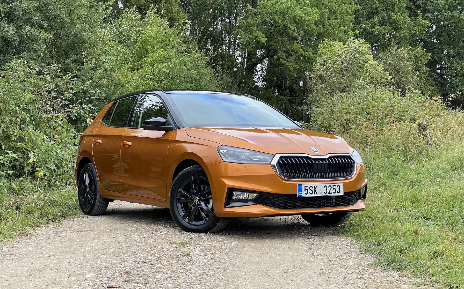 Full sidoprofil av en modern, orangefärgad Skoda Fabia 2021 halvkombi parkerad på en grusväg med växtlighet i bakgrunden, tagen från en något sned vinkel framifrån som framhäver förarsidan. Funktioner inkluderar en framträdande grill med centrerad logotyp, eleganta strålkastare, dimljus, stora svarta flerspokedade lättmetallfälgar, karossfärgade dörrhandtag och speglar, sluttande taklinje och en antydan till den bakre luckan och ljusenheten.