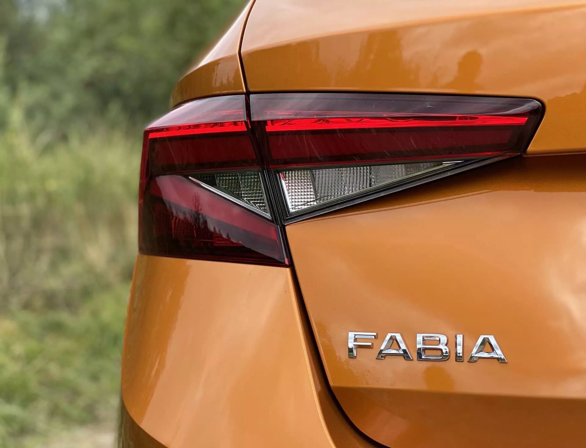 Närbild av det bakre hörnet på en 2021 Skoda Fabia som lyfter fram den skarpa, kantiga designen på bakre LED-ljussammansättning och den distinkta 'FABIA'-logotypen på den glansiga, metalliska orangefärgade karossen. De mjuka kurvorna och försiktiga buktningarna runt baklyktan bidrar till den moderna estetiken, fångad från en något upphöjd vinkel med liten skärpedjup.