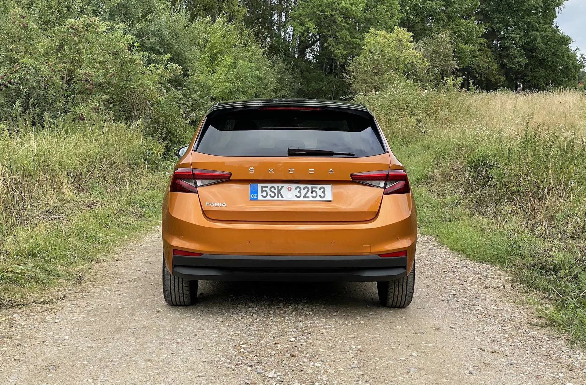 Bakre vy av en ljus metallisk orange Skoda Fabia 2021 hatchback fotograferad i dagsljus på en smal grusväg i en lantlig miljö. Bilden fångar den moderna bakre designen med en kontinuerlig bakljuslist, vassa vinklade bakljus, integrerade reflektorer, bakruta med torkare och en bakspoiler för aerodynamik, vilket framhäver bilens glansiga yta och praktiska funktioner.