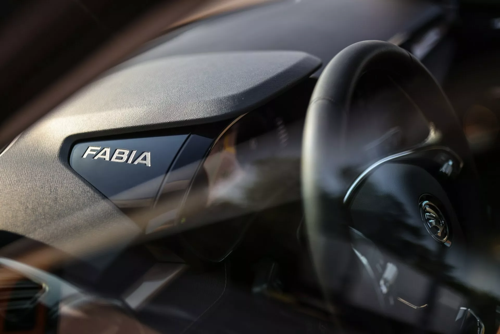 Närbild av interiören i en Skoda Fabia 2021, med fokus på passagerarsidan av instrumentbrädan. Ordet 'FABIA' visas tydligt, medan den moderna ratten med Skoda-logotypen är delvis synlig till vänster. Instrumentbrädan har en kornig struktur och det grunda skärpedjupet betonar varumärket med en mjukt oskarp bakgrund.