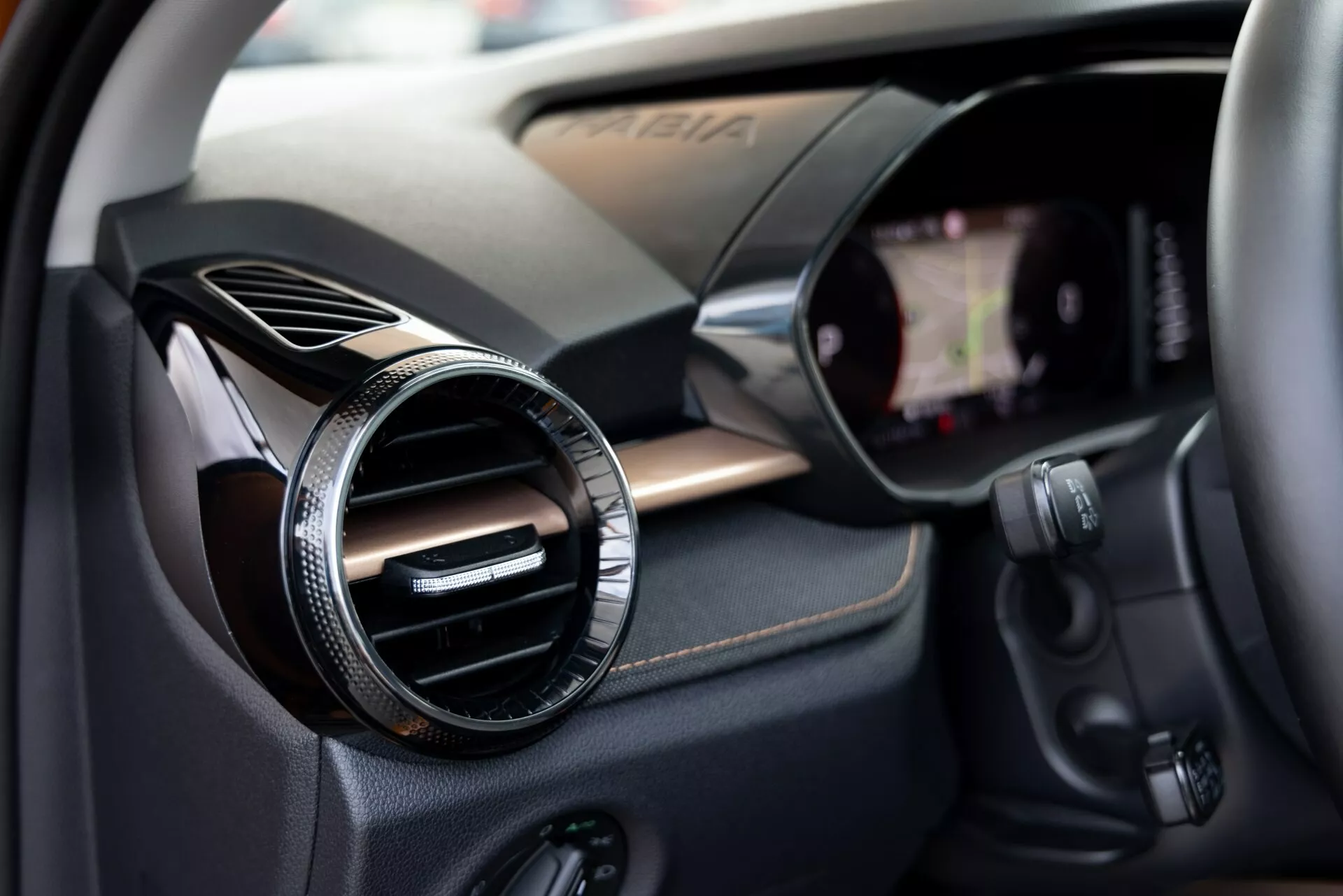 Närbild av interiören i Škoda Fabia 2021 fokuserad på passagerarsidan. Bilden visar en rund luftventil med en metall- eller kromfinish mot en kolfiberliknande texturerad panel, vilket indikerar en sportig eller lyxig trimnivå. Till höger är en kant av pekskärmen för infotainmentsystemet integrerad i instrumentbrädan synlig. Under luftventilen finns olika kontrollknappar, möjligen för ljudsystemet, klimatkontrollen eller andra fordonsegenskaper. Det grunda skärpedjupet drar uppmärksamheten till luftventilen samtidigt som det mjukt antyder de omgivande teknologiska komponenterna.