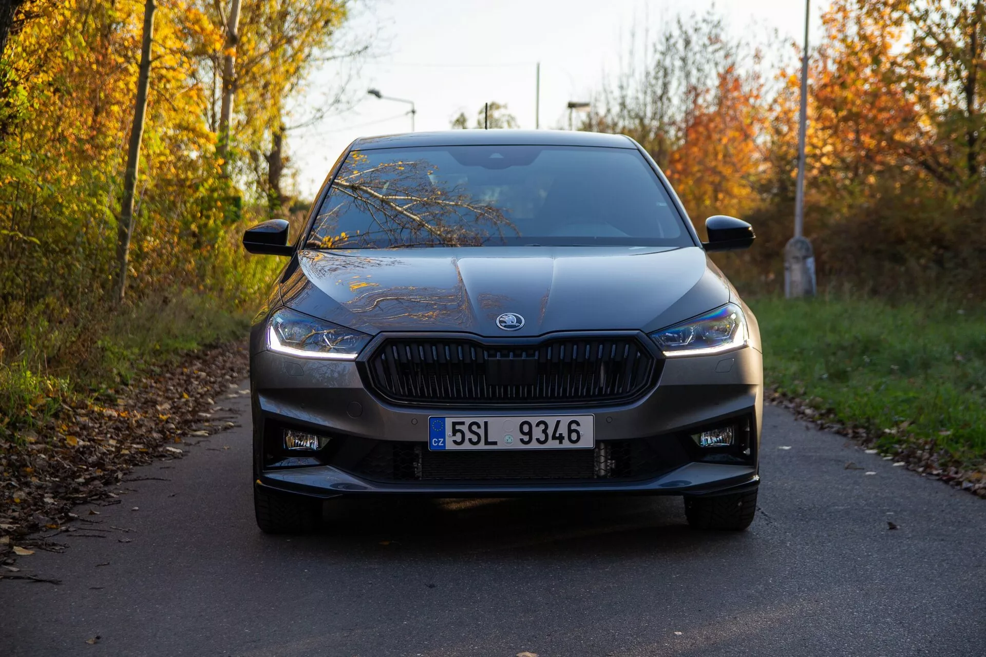 Frontal vy av en Skoda Fabia 2021 tagen från låg vinkel, vilket framhäver det distinkta grillningsröret med vertikala spjälor och moderna, smala strålkastare. Bilen, centrerad i ramen, är parkerad på en smal väg kantad av höstlöv, med tillverkarens emblem tydligt synligt i centrum av grillen. Ytterligare design detaljer som är synliga inkluderar den tydliga registreringsskylten, luftintagen på nedre stötfångaren, mjuka konturlinjer på huven, expansiv vindruta och sidobackspeglar med indikatorer. Den mjuka belysningen och de långa skuggorna tyder på att bilden togs antingen tidigt på morgonen eller sent på eftermiddagen, och visar bilen i utmärkt skick.