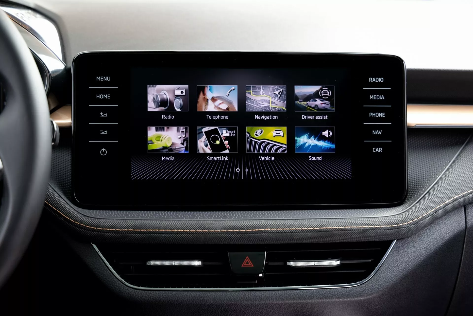 Närbild av infotainmentskärmen i 2021 Škoda Fabia, fotograferad från framsätet antingen som passagerare eller förare. Mittkonsolen visar en elegant design som troligen är inramad av borstad aluminium. Skärmen visar ett användarvänligt grafiskt gränssnitt med färgglada ikoner för Hem, Radio, Telefon, Navigation och mer. Under skärmen finns en rad med fysiska knappar och en möjlig CD-spår, vilket understryker multifunktionaliteten. Premiumklädsel med kontrasterande sömmar antyder en exklusiv interiördesign.