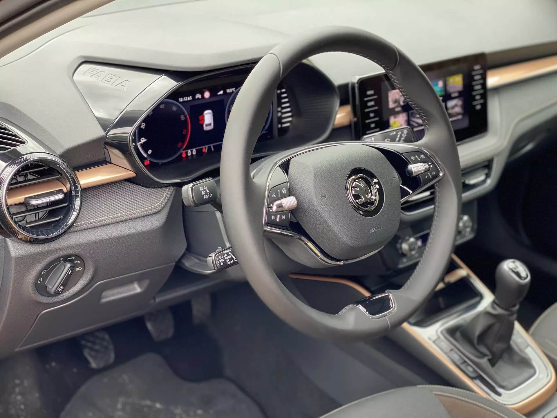 Interiörvy av Skoda Fabia 2021 fokuserad på förarsidan, som visar den centrerade ratten med multifunktionella kontroller för ljud, röstkommandon och farthållare. Den digitala instrumentpanelen visar hastighetsmätare, varvräknare och fordonsstatusindikatorer. Ett pekskärmsinfotainmentsystem visas på instrumentbrädan med navigations- och mediefunktioner. Under skärmen finns klimatkontrollknappar och rattar. En manuell växelspak är synlig på mittkonsolen.