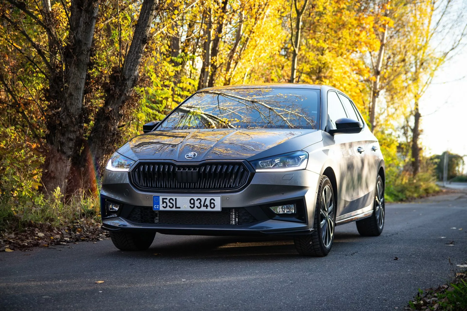 Bild på en modern Skoda Fabia 2021 sedan parkerad på en väg kantad av höstlövverk, tagen från ett markperspektiv, som framhäver den främre grillen, vinklade strålkastare, eleganta designlinjer, lättmetallfälgar och sidospeglar med integrerade blinkers, som speglar en högkvalitativ metallfinish och en kombination av lyx och sportighet.