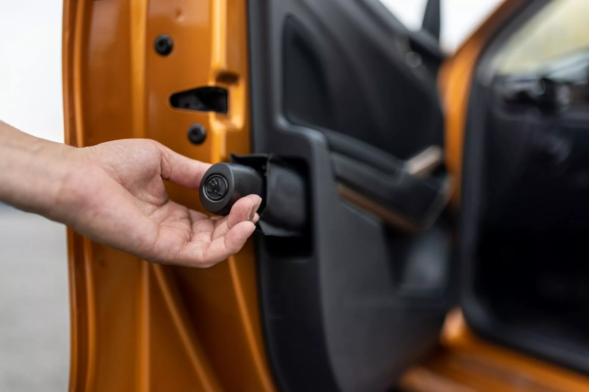 Närbild av förardörren på Skoda Fabia 2021, något på glänt, sedd från utsidan med den inre kanten synlig. En hand håller en kompakt, svart cirkulär nyckelbricka som indikerar ett nyckelfritt system eller bilsäkerhetssystem. Bilen är ljusorange med en metallisk finish och visar avancerade låsmekanismer och anpassade säkerhetsfunktioner.
