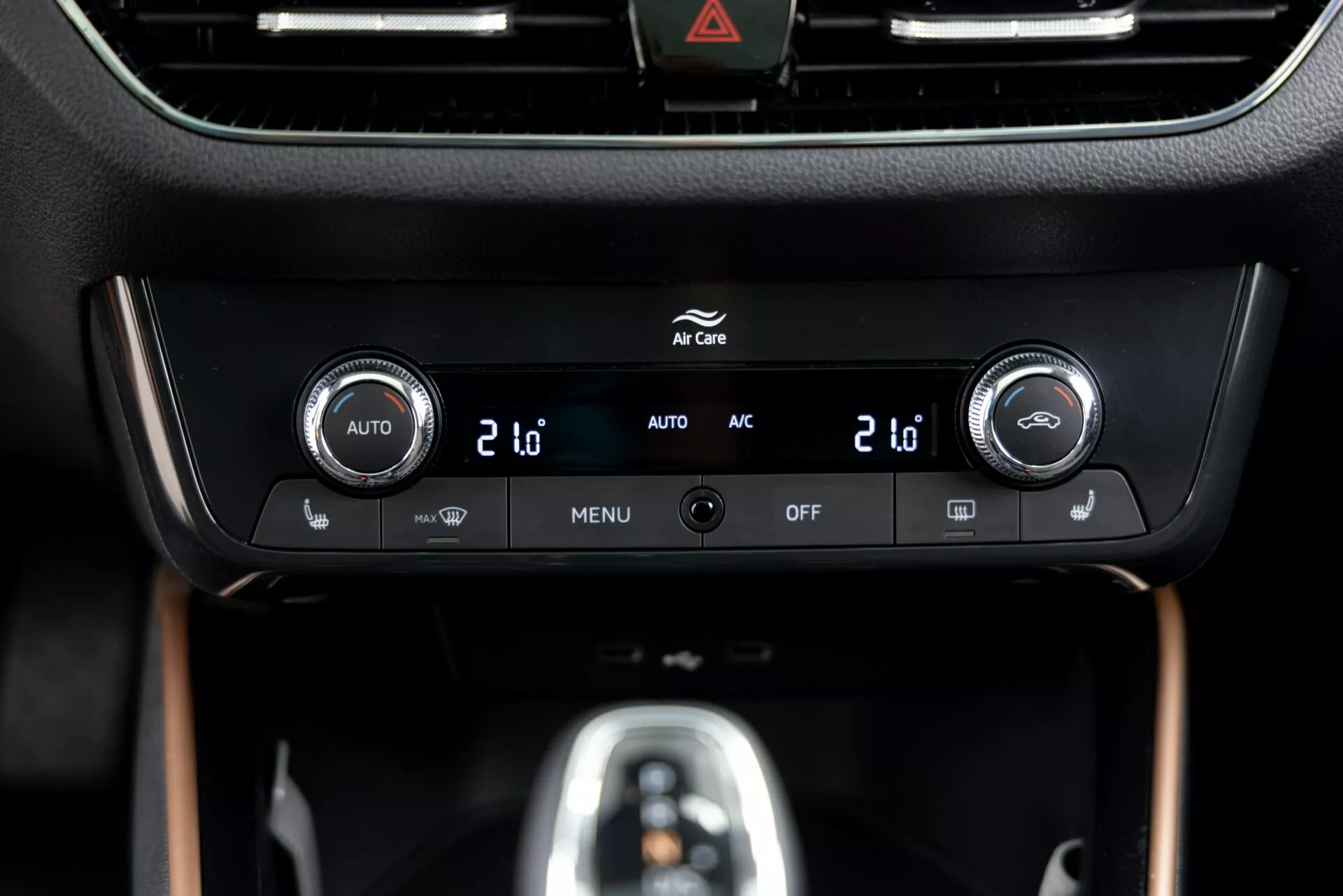 Närbild av klimatkontrollpanelen på Skoda Fabia 2021, placerad på mittkonsolen, med dubbla temperaturreglage med digitala skärmar som visar '20', integrerade knappar för automatisk klimatanläggning ('AUTO'), luftkonditionering ('AC'), menyinställningar ('MENU') och systemavstängning ('OFF'). En glansig svart ram omger en informationsskärm ovanför kontrollerna, med en synlig varningsknapp (triangelsymbol) överst. Panelen är inbäddad i en premium inredning med matt och träliknande ytor, vilket framhäver en modern design och teknik.