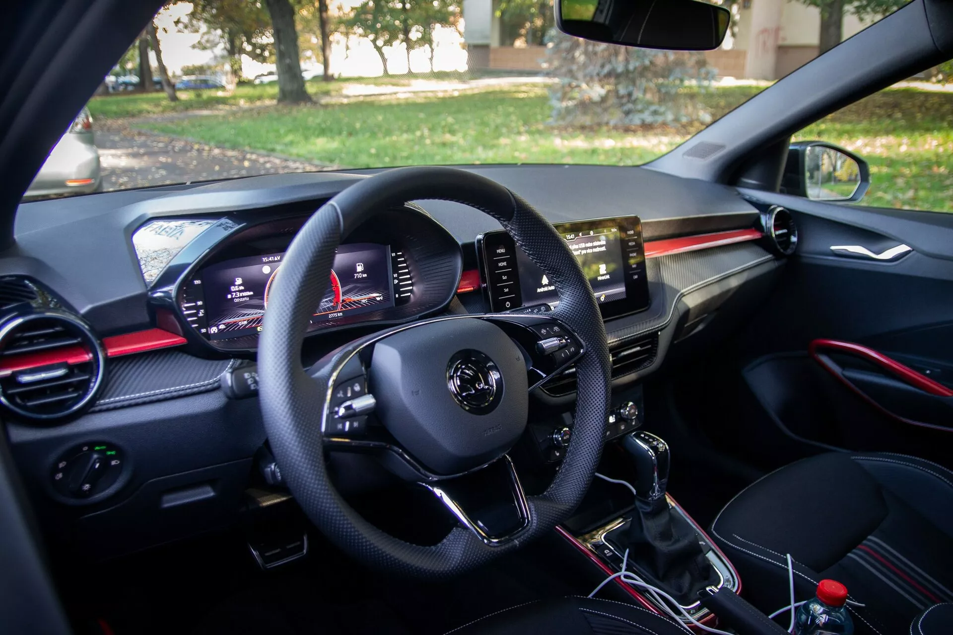 Interiörvy av en modern 2021 Skoda Fabia från förarens perspektiv, med kontrollpanel på förardörren, multifunktionell ratt, digital instrumentpanel, luftventiler med röda accenter, central infotainment pekskärm, manuell växelspak och passagerarsätesområdet med handskfack och matchande luftventil.