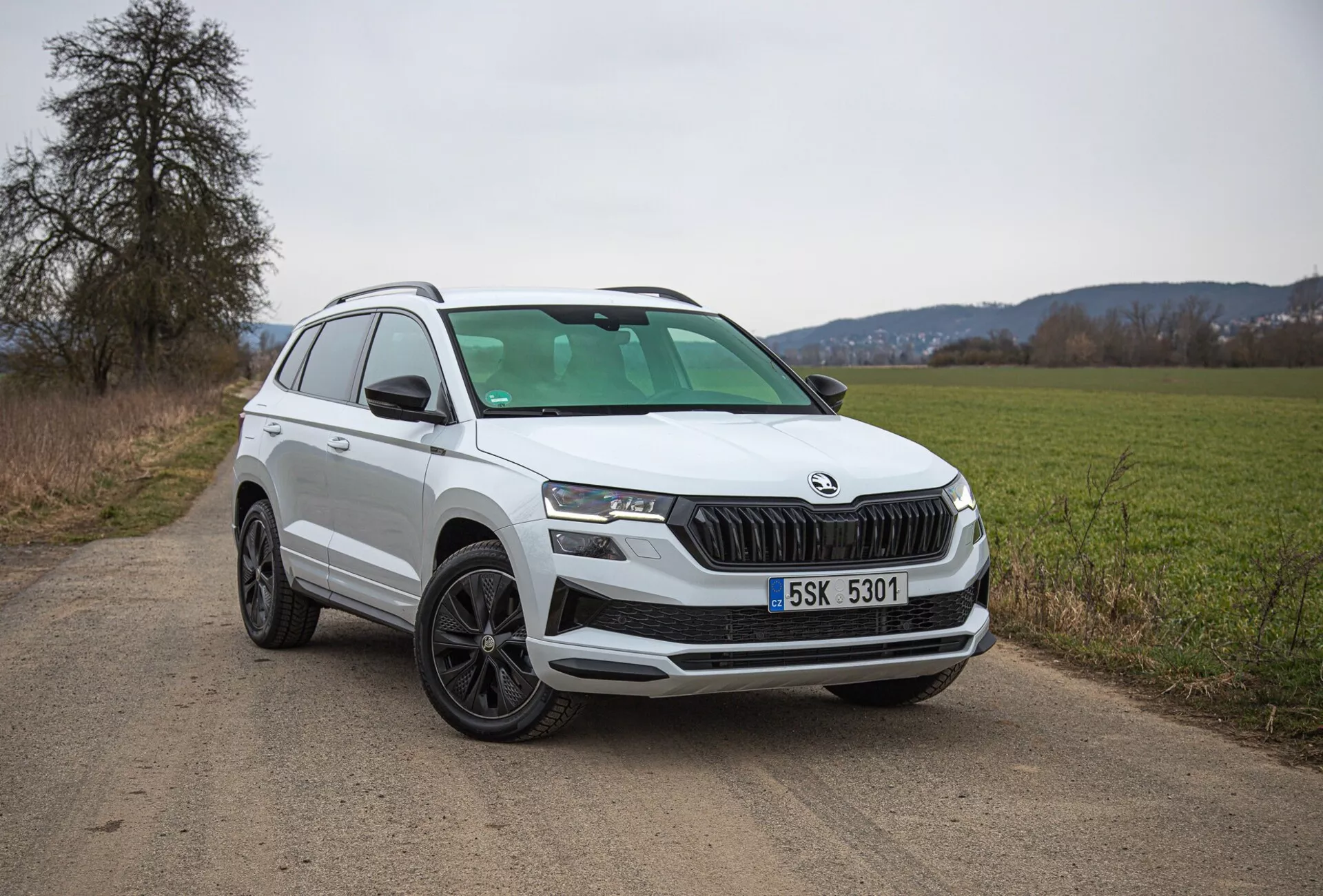 Vit SUV Škoda Karoq 2021 parkerad på en landsväg av hårt packad jord, visar framifrån vänster kvartalvy inklusive grill, strålkastare och hjuldesign, med fokus på moderna funktioner som LED-belysning och parkeringssensorer.