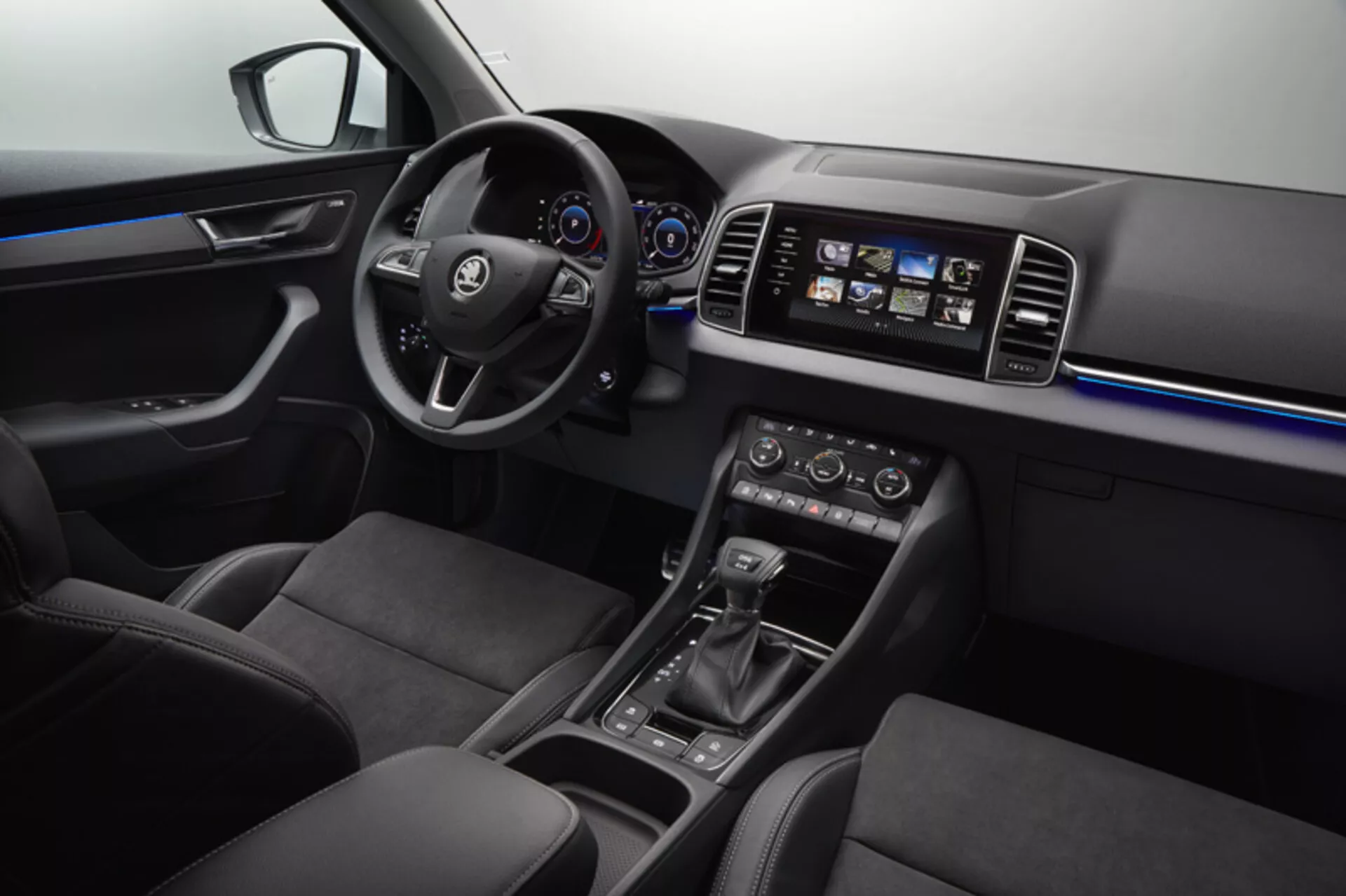 Interiörvy av Skoda Karoq 2017 som visar instrumentbrädan, förardörren med kontroller för fönster och speglar, ratten med multimedia- och farthållarknappar, digital-analog instrumentkluster, pekskärms infotainmentsystem, dubbelzons klimatkontroll, automatisk växelväljare, val av körningsläge och säten klädda i läder