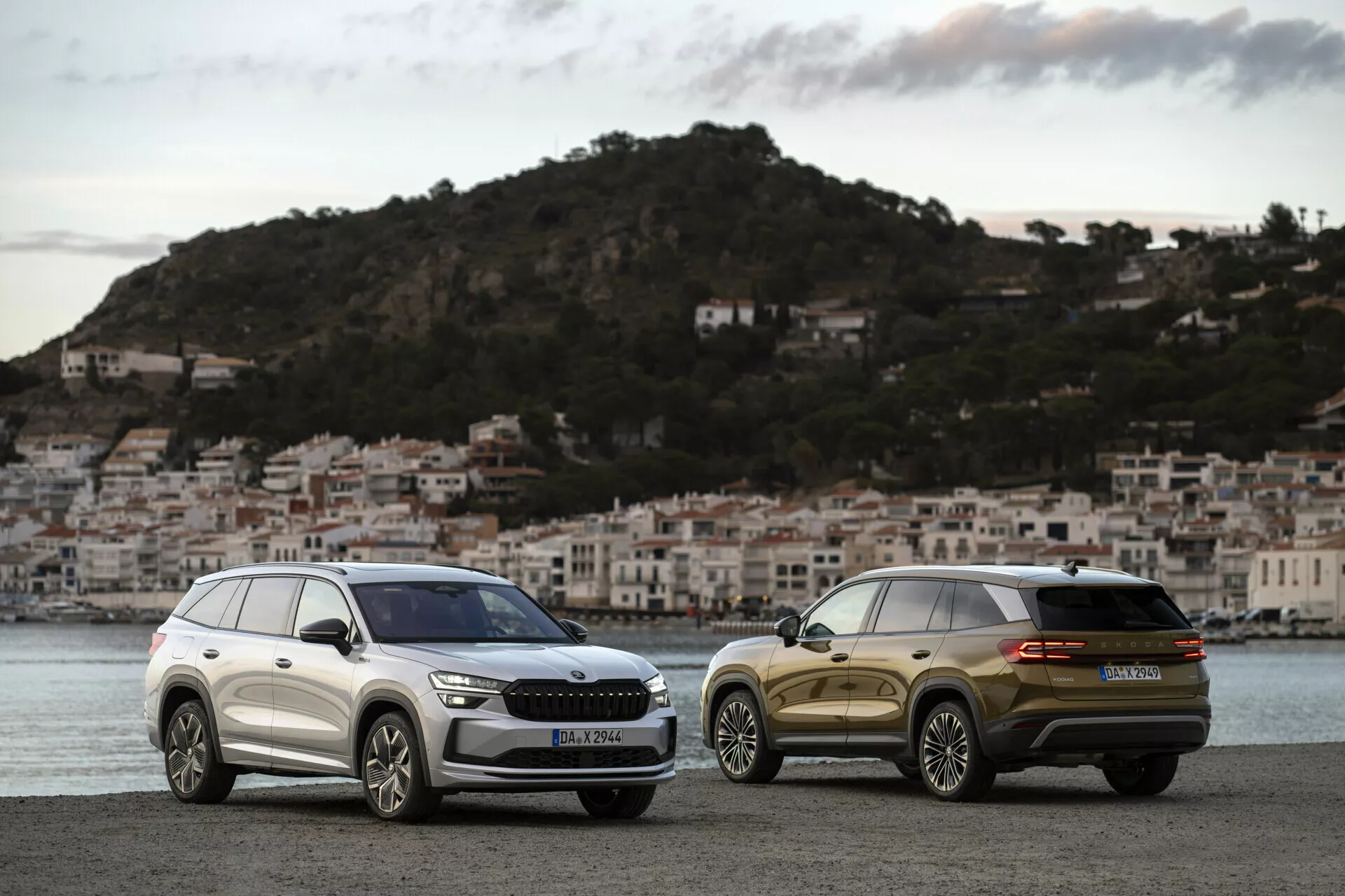 SUV Skoda Kodiaq 2024 parkerade sida vid sida i gryningen på en strandpromenad med en kuststad i bakgrunden, som visar den främre grillen, strålkastarna och de eleganta designlinjerna för den ena och den bakre trekvartsutsikten för den andra.