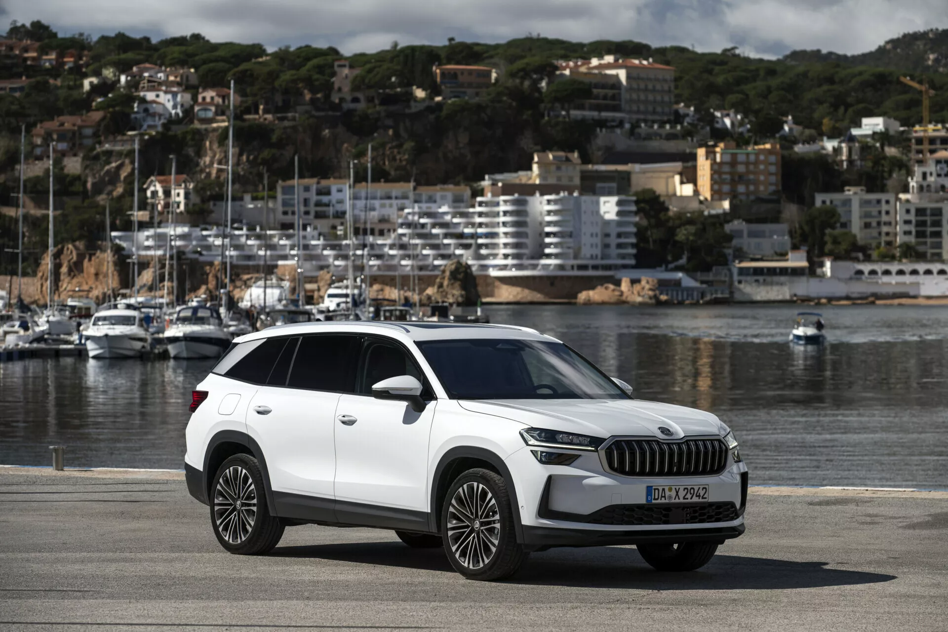 2024 Skoda Kodiaq crossover SUV parkerad vid en havsnära promenad som visar hela bilens vy inklusive moderna designelement som LED-strålkastare, stor grill, flerspakiga lättmetallfälgar, med scenisk båt och urban bakgrund