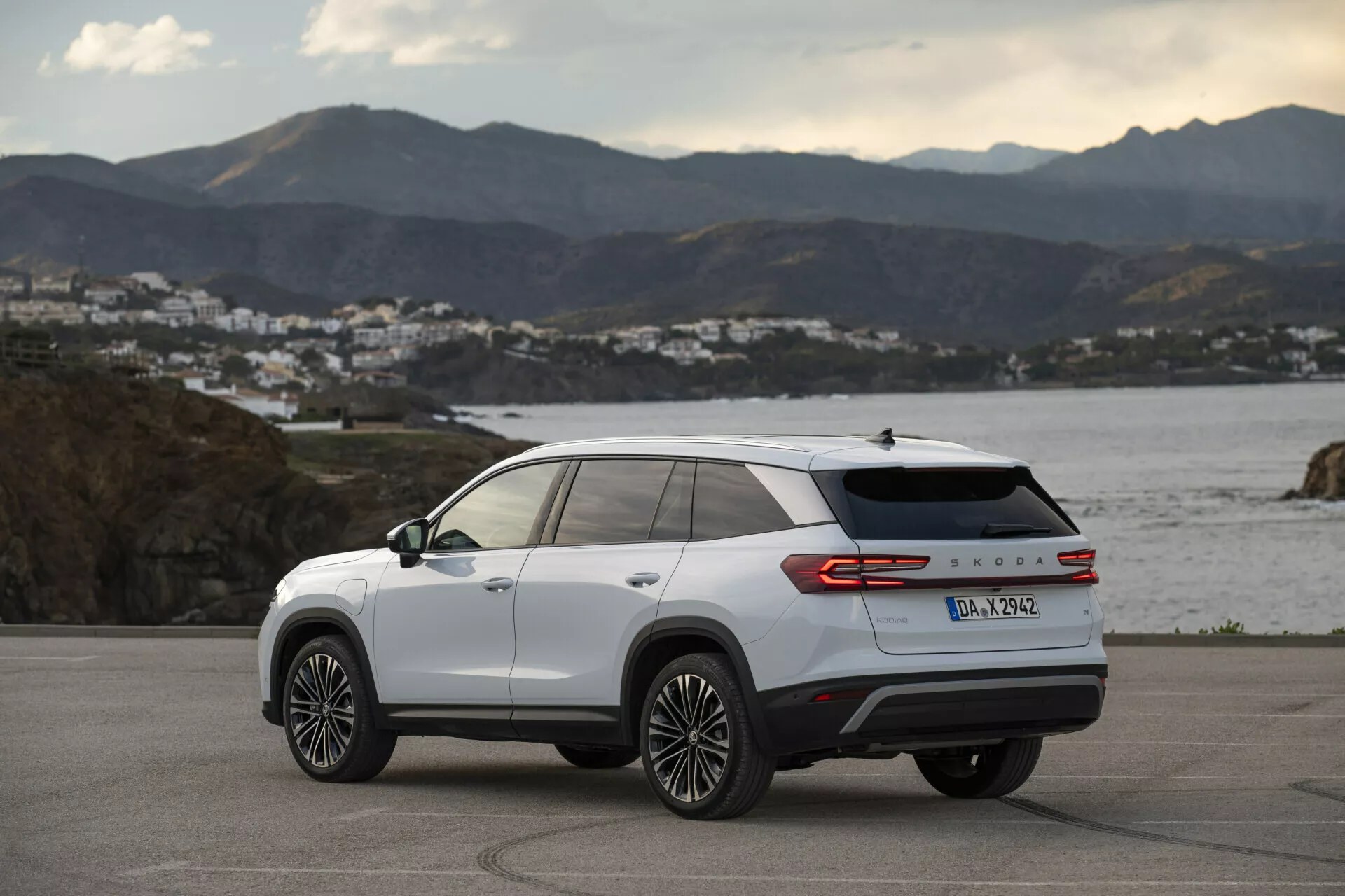Trekvarts bakifrån vy av en vit Skoda Kodiaq 2024, som visar sidprofil och baktill designlinjer, flerspakiga aluminiumsfälgar, LED-baklampor och modellbeteckning, parkerad utomhus på en kustväg vid gyllene timmen