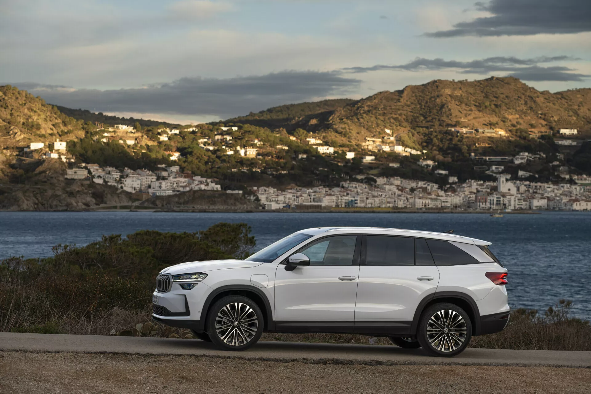 Modern SUV Skoda Kodiaq 2024 parkerad längs en kustväg med en naturskön utsikt över en bukt och hus på kullarna i bakgrunden, med en slank design, lång hjulbas, hög markfrigång, skarpa karosserilinjer, inåtgående dörrhandtag, tonade bakre fönster, takräcken, stora legeringshjul, LED-strålkastare, sidospeglar med integrerade blinkers och ett panoramasoltak, allt mot en kontrasterande vit exteriörfinish.