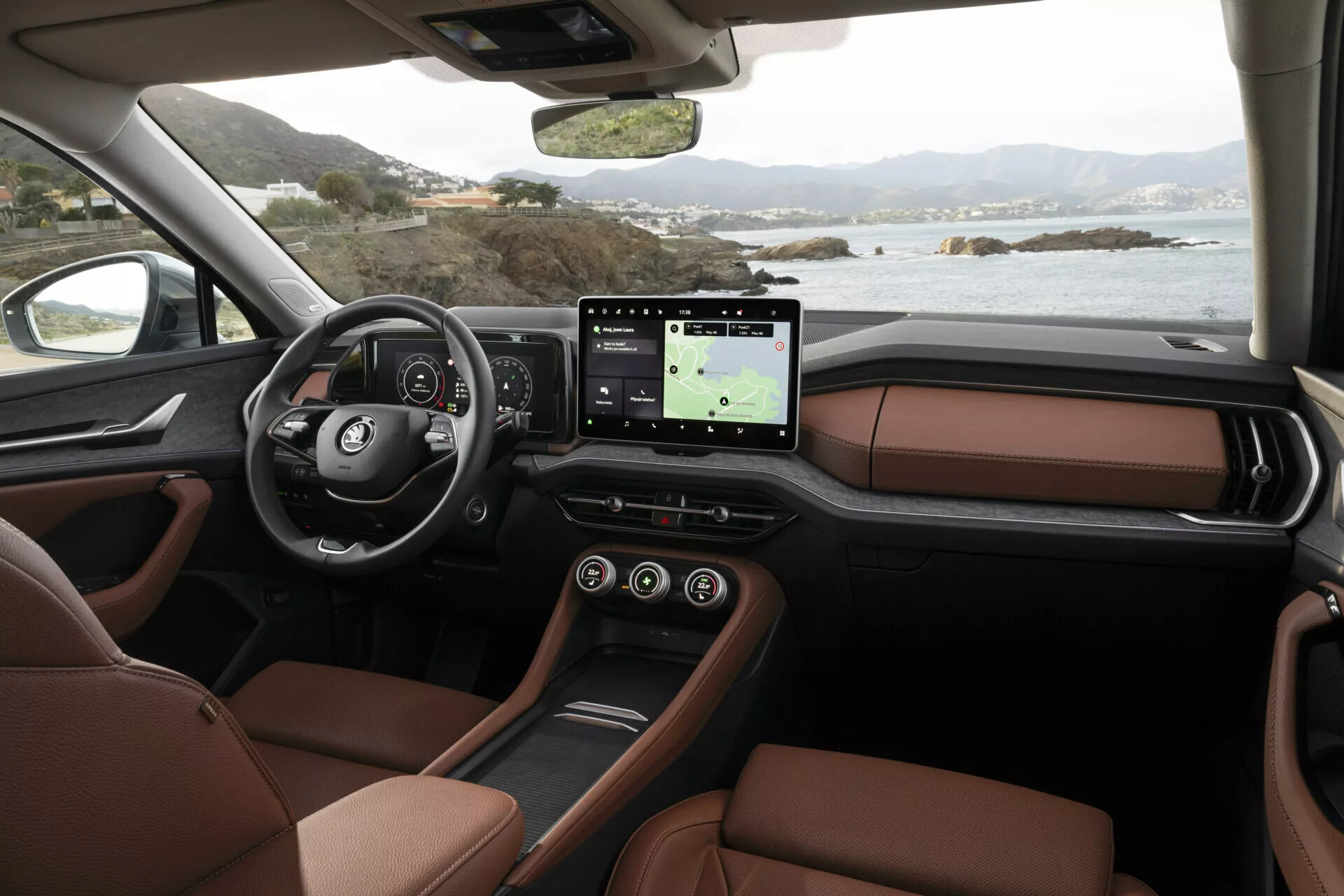 Interiörvy av Skoda Kodiaq 2024 från passagerarperspektiv som visar instrumentbrädan, multifunktionsratt med logotyp, digital instrumentkluster, stort pekskärmsinfotainmentsystem med GPS, lyxiga bruna lädersäten och en elegant mittkonsoldesign, mot en fridfull kustlandskap utanför fönstren