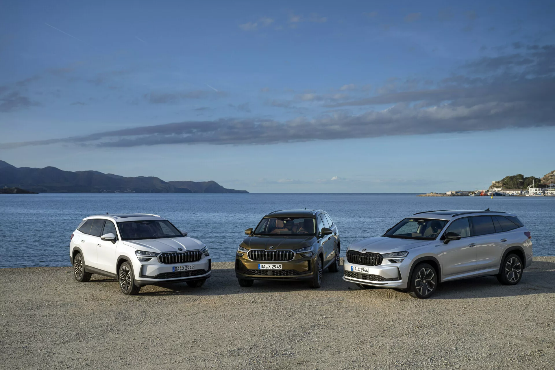 2024 års Skoda Kodiaq SUV-sortiment i vit och mörk färg i en främre sidovy, parkerad vid en vattenkant med berg och delvis molnig himmel i bakgrunden