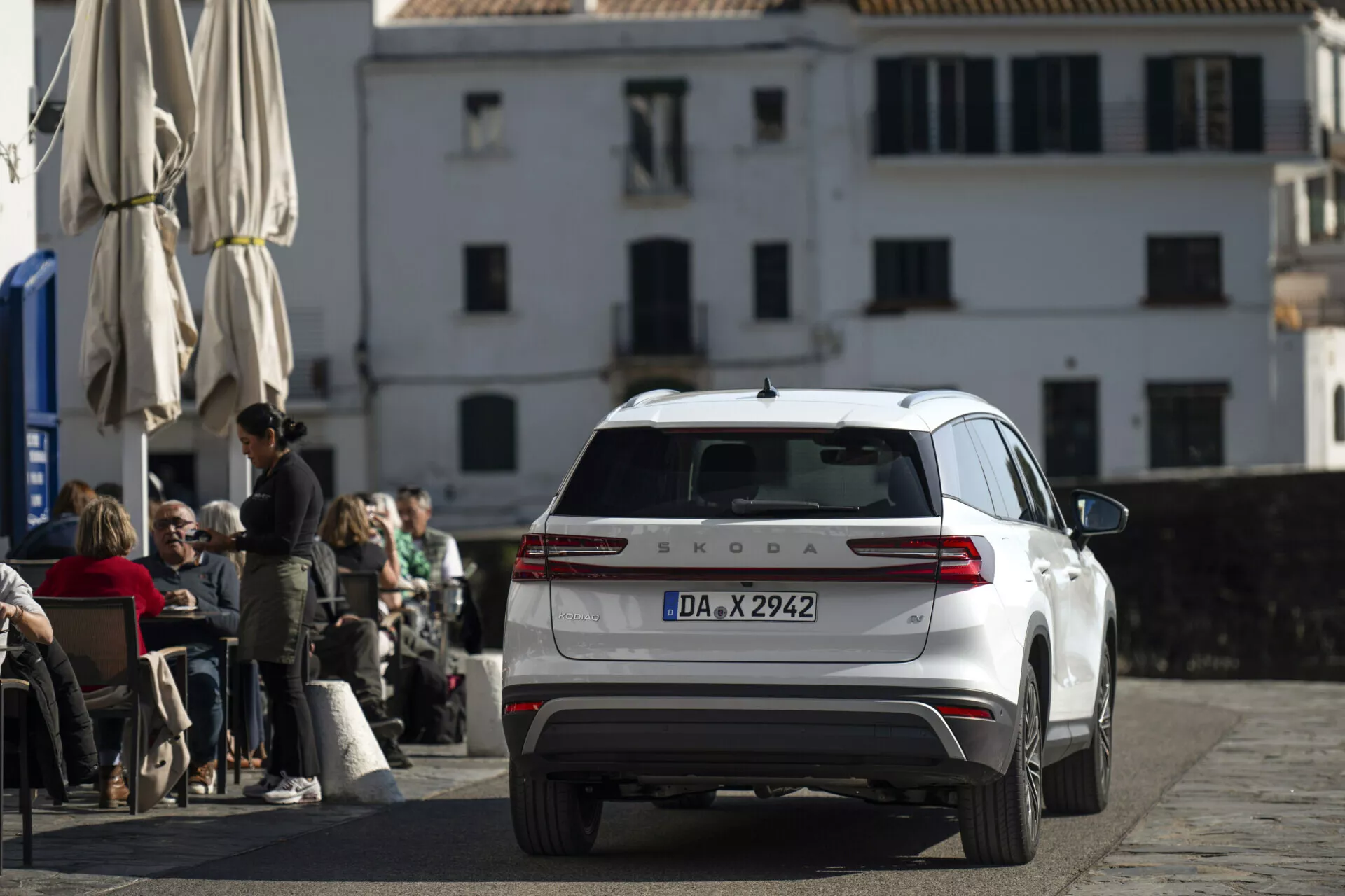 Vit Škoda Kodiaq 2024 fotograferad från ett bakre, något upphöjt vinkel, visar hela baksidan, bakljus, bagageluckan, bakfönster med torkare och tak med antenn och räcken, parkerad på en kullerstensgata framför ett café med klassisk europeisk arkitektur i bakgrunden.
